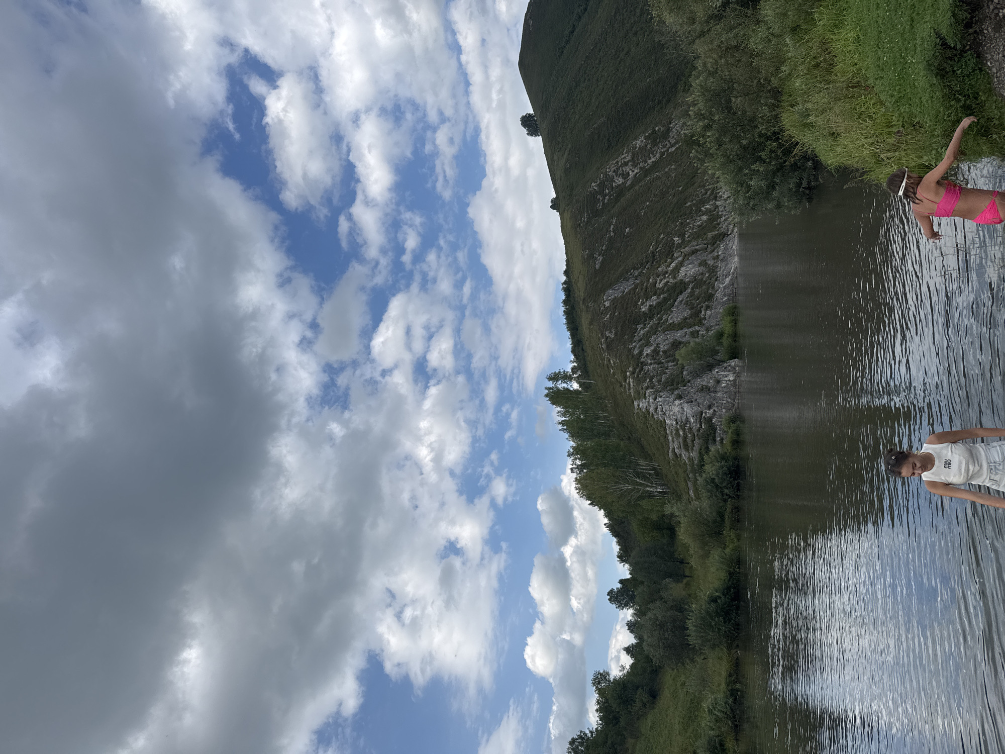 This media file depicts a scenic landscape with two young individuals by a body of water. The scene is oriented vertically, with the sky occupying the left side and the landscape and water on the right.
The sky is filled with a mix of large, fluffy white clouds and some darker, grayer clouds, suggesting a partly cloudy day. Patches of blue sky are visible between the clouds. A steep, grassy hill slopes down towards the water on the right side of the image. The hill is covered in dense green grass and scattered trees, with a rocky, lighter-colored section visible higher up.
In the foreground on the right, a young person in a bright pink bikini stands at the edge of the water, looking down and to the left. They are wearing a white headband. Further to the right and slightly behind, another young person in a white tank top and shorts is visible. Both appear to be at a calm, reflective body of water, possibly a river or lake. The water's surface is rippled, reflecting the sky and surrounding greenery. The overall atmosphere seems relaxed and summery. The presence of the grassy hill and the type of vegetation suggest a natural, possibly rural or mountainous environment. The time of day appears to be daytime, with ample natural light.
There is no visible text in the image. The location is identified as Soloneshnoye, Russia.