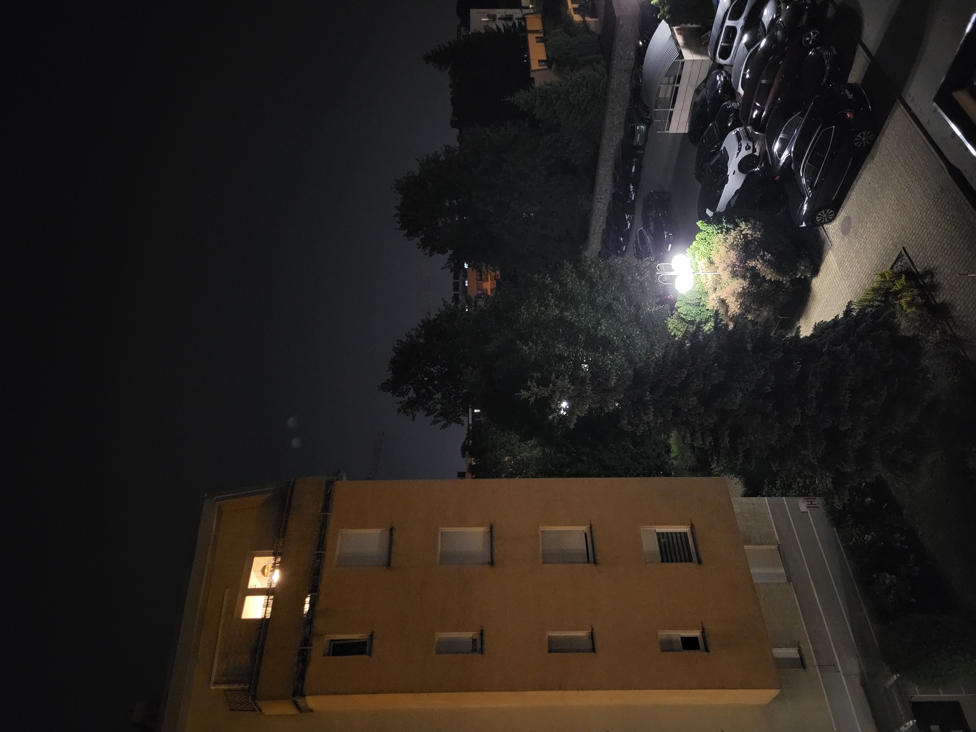 The image depicts a night scene in an urban environment. A multi-story building with several windows is in the foreground. Some windows are lit, indicating occupancy. Above the building, trees fill the space, with a streetlamp illuminating a section of the foliage. A parking area with several cars is visible to the right, and another building is in the background. The sky is dark, with a few indistinct light sources visible.