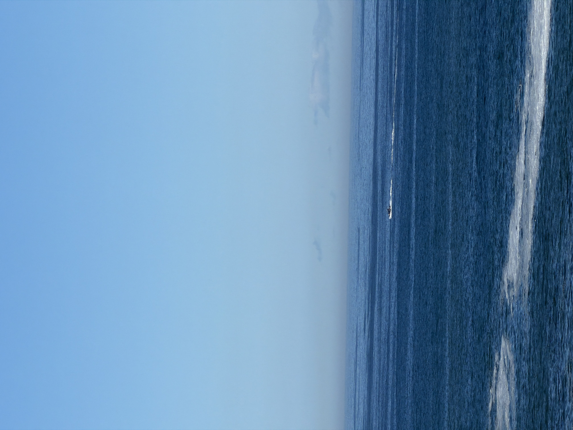 The image shows a seascape with a clear blue sky occupying a significant portion of the left side of the frame. The ocean, a deep blue, fills the right side. Ripples and streaks of white foam indicate the presence of waves or currents. A small, dark shape, possibly a boat or jet ski, is visible on the water's surface. Given the presence of the ocean and the clear sky, the location is likely coastal. Without additional landmarks or information, it is difficult to specifically pinpoint the exact location within Cape Town, South Africa. The scene depicts a marine environment with water and sky as the dominant elements. A single vessel is active on the water.
