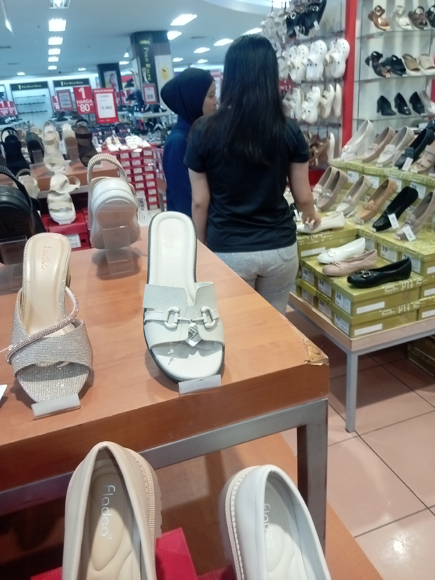 The image was taken inside a shoe store in Batam, Indonesia. The store features rows of shoes displayed on shelves and tables. In the foreground, there are several pairs of women's shoes, including heels and flats. Two women are visible in the image. One woman, wearing a black headscarf and a blue top, is standing and looking towards the shoes. Another woman, with long dark hair, is wearing a black t-shirt and light grey pants, and is also looking at the shoes. The store appears to be well-lit with fluorescent lighting.