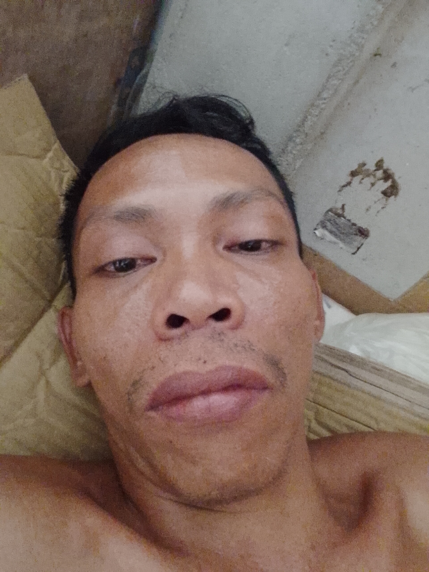 A close-up selfie of a man with dark hair, taken indoors. His face is oily and reflects light. He has a slight mustache. Behind him are a cardboard box, a section of a white-painted wall with patches and debris, and white bedding. It is not possible to determine the exact location in Bukit Ketok from the image alone.
