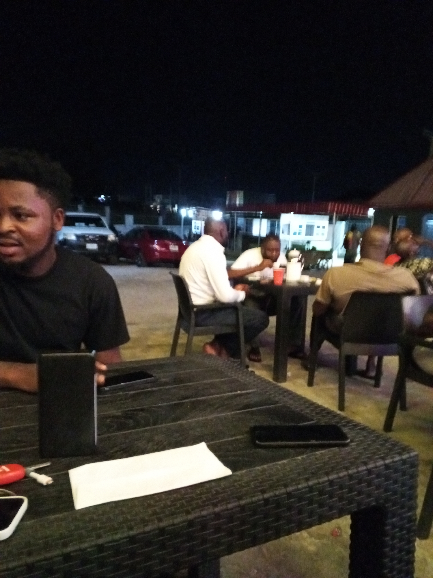 The image captures an outdoor evening scene in Lokoja, Nigeria. A group of men is seated at tables, engaged in conversation and eating. In the foreground, a man with short hair, wearing a black t-shirt, is visible at the corner of the table. Various items such as phones, a wallet, and a tissue pack are present on the table. Behind the foreground table, other men are seated at two other tables. A man in a white shirt sits at the center table, while another man in a brown shirt is seen seated on the table to the right. Several cars can be seen parked in the background, along with low buildings. The scene is lit by artificial lights.