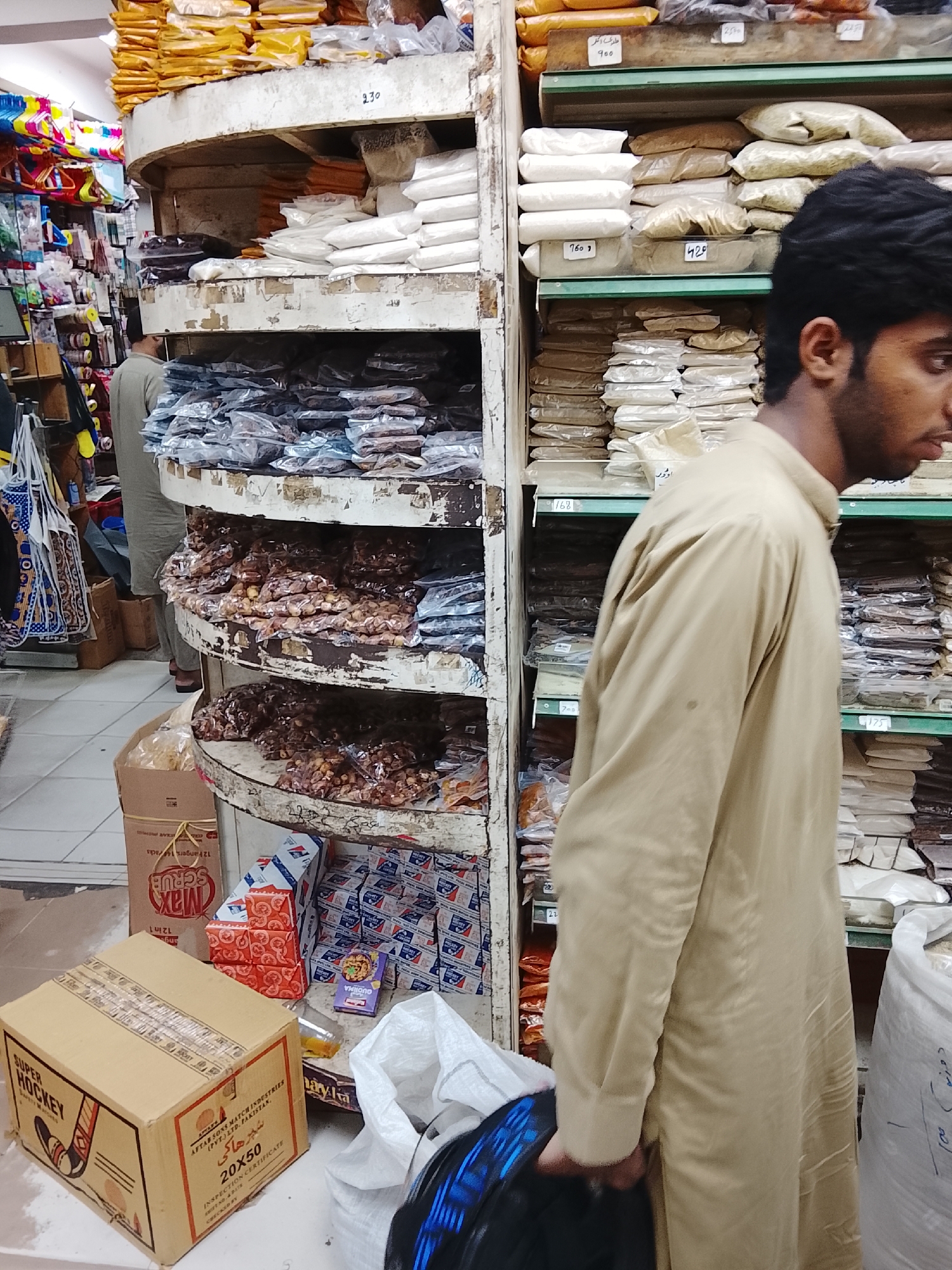 The image shows the interior of a small grocery store in Gulgasht Colony, Pakistan. A man wearing a light brown kurta is standing in the right foreground with his face partially visible. He appears to be a customer. Behind him, to the left, is a circular shelving unit stocked with various bagged goods, likely food items such as spices, nuts, and flour. The shelves are somewhat worn and have handwritten price tags. To the left of the shelving unit, there are items such as hanging toys, bags, and more stacked goods. In the left foreground, there are boxes of "Super Hockey" matches. Additional boxed and packaged items are on the floor near the shelving unit. Another man is further back in the store. The store appears crowded and typical of a small retail establishment.