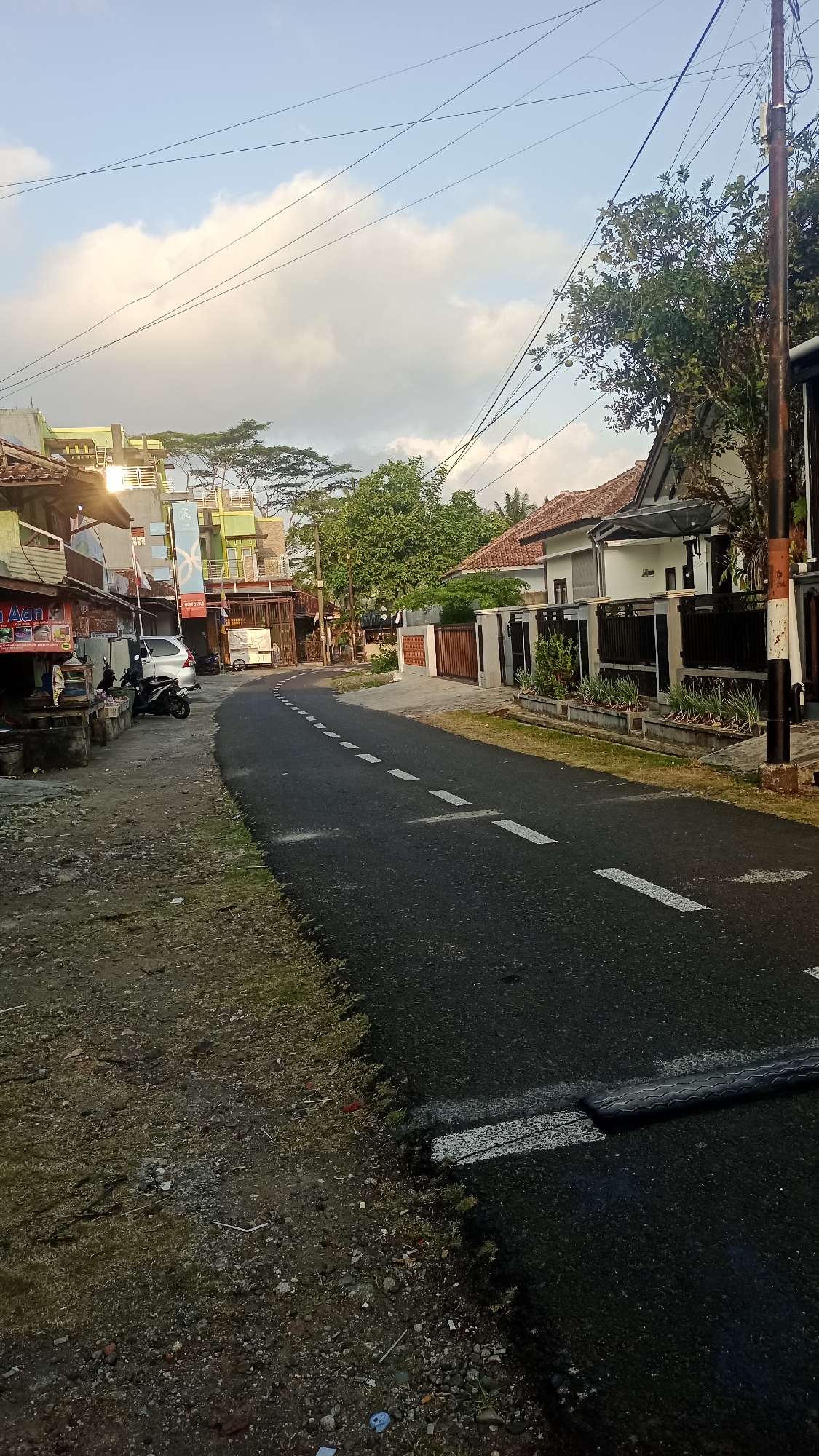 The image depicts a street scene in Pakemitan, Indonesia. The road, paved with asphalt, features a dashed white line marking the center. On the left side of the road, a small roadside business is visible, with a motorcycle parked in front of it. Further down the road on the left, a silver car is parked. Buildings line the street, some appearing to be residential homes with terracotta roofs and modern fences, while others seem to be commercial establishments with multiple stories and bright paint colors. Trees and vegetation are scattered throughout the scene, with lush green foliage evident. The sky is partly cloudy with a bright, diffused light, suggesting it is either early morning or late afternoon. The overall environment appears to be a typical street in a semi-urban or rural Indonesian setting, with a mix of residential and commercial properties.