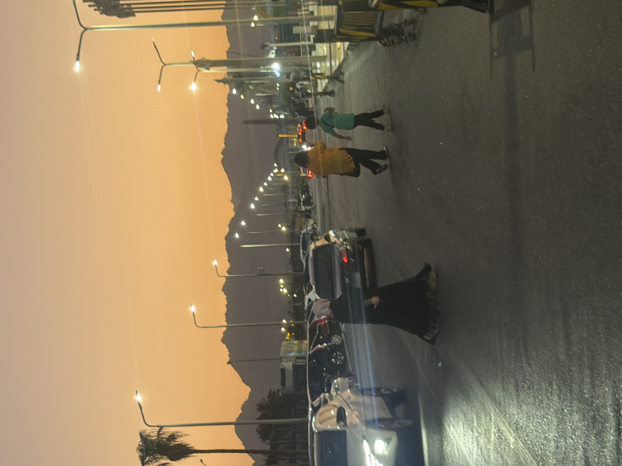 The image captures a street scene in Sharm El Sheikh, Egypt, during dusk. The sky is a prominent feature, painted in warm hues of orange and yellow, characteristic of a sunset. Silhouetted against the sky are distant mountains, adding a dramatic backdrop to the urban environment.
In the foreground, a paved road is visible, lined with streetlights that are illuminated, casting a soft glow. Several vehicles are parked or driving on the road, suggesting a moderately busy street. Among the vehicles, a white SUV is prominent on the left side of the frame, its headlights on.
Two people are walking across the road. One is a person wearing a black abaya and hijab, their face obscured. They are walking from right to left across the frame. To their right and slightly ahead, a woman in a bright yellow top and dark pants is walking, accompanied by a young boy in a green t-shirt and dark pants. They appear to be walking in the same direction as the person in the abaya.
Further back, more cars and streetlights are visible, along with some palm trees on the left side, indicating a warm climate. The overall impression is one of a typical evening scene in a coastal city, with a blend of natural beauty and urban activity.