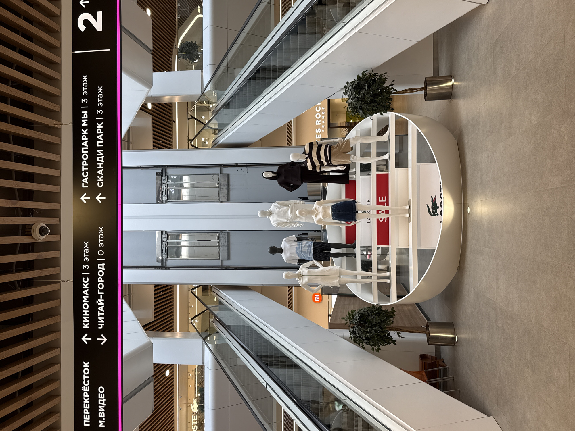 The image depicts the interior of a modern shopping mall, likely in Izhevsk, Russia, given the Cyrillic signage. The scene is dominated by escalators on either side, leading upwards. In the center, a multi-tiered display features mannequins dressed in casual clothing, including a woman in a white zipped dress and denim skirt, a man in a t-shirt and shorts, and another man in a striped shirt and shorts. One mannequin is wearing a black hooded garment. Below the mannequins, a large red "SALE" sign is visible, along with a "LACOSTE" logo.
Above the central display, prominent signage indicates directions to various sections of the mall. On the left, "ПЕРЕКРЁСТОК" (Crossroads) and "М.ВИДЕО" (M.Video, an electronics retailer) are indicated, with arrows pointing to "КИНОМАКС | 3 этаж" (Kinamax | 3rd floor) and "ЧИТАЙ-ГОРОД | 0 этаж" (Chitai-Gorod | 0th floor, a bookstore). On the right, signs point to "ГАСТРОПАРК МЫ | 3 этаж" (GastroPark My | 3rd floor) and "СКАНДИ ПАРК | 3 этаж" (Scandi Park | 3rd floor). A large number "2" with an upward arrow is also displayed on the right.
The mall's architecture features clean lines, white surfaces, and wood-paneled ceilings. Plants in metal planters are positioned near the escalators and the central display, adding a touch of greenery. In the background, shopfronts with illuminated signs are visible, including "LACOSTE" and "YVES ROCHER FRANCE." The floor is tiled in a neutral tone. A security camera is mounted on the ceiling in the upper left portion of the image. There are no identifiable people actively shopping or interacting in the scene, suggesting it was photographed during a quiet period or from a distance.