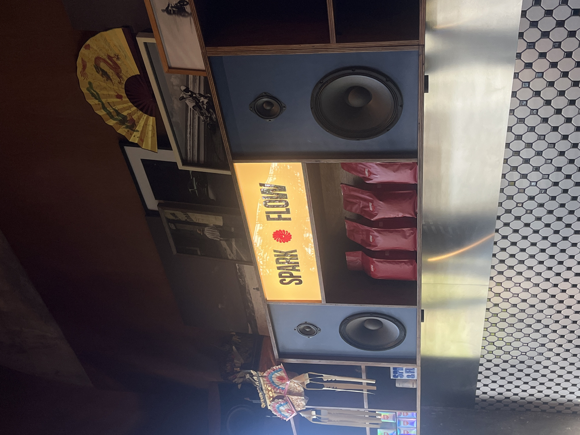 The image depicts a shelf or display in North Kuta, Indonesia, featuring a "Spark Flow" sign illuminated with a warm light. There are two large speakers mounted on blue panels, one above and one below the sign. Between the sign and the speaker above are several red "Campus" branded items, seemingly stacked. Next to the speaker panel is a reflective surface, adjacent to a wall covered in black and white geometric patterned tiles. To the left of the speaker structure are framed photographs and an ornamental fan. Below the lower speaker are various items including a decorative parasol and some printed materials. The background fades into darkness, suggesting the surrounding environment is dimly lit.