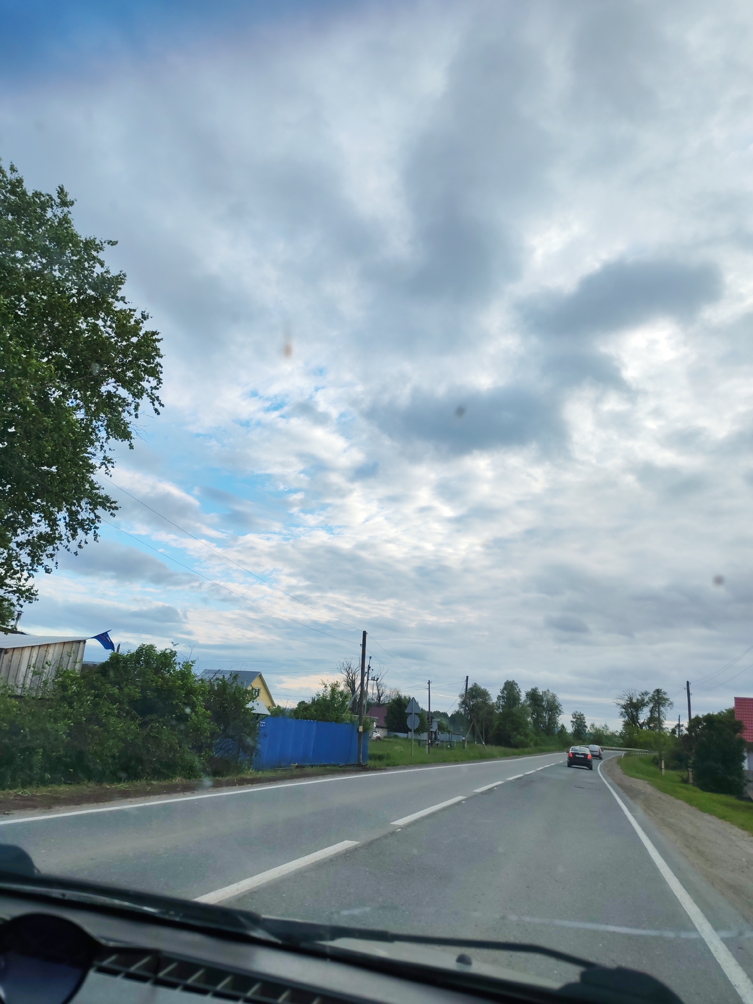 The image captures a view from inside a vehicle looking forward through the windshield onto a rural road in Malaya Bod'ya, Russia. The bottom portion of the image shows the vehicle's dashboard and the lower edge of the windshield, with several dark, circular smudges indicating reflections or dirt on the glass. The two-lane asphalt road extends into the distance, curving slightly to the right, with a solid white line delineating the right shoulder and a dashed white line separating traffic directions. Two passenger vehicles are visible ahead, traveling in the same direction; the lead car is dark-colored, followed by a lighter-colored vehicle.
On the left side of the road, a dense green tree partially obstructs the upper-left sky. Beneath the tree and alongside the road are various bushes and utility poles with overhead wires. A wooden building and a vibrant blue corrugated metal fence are visible, separating properties from the roadside. Further along the left, another house with a yellow exterior wall is partially visible. The right side of the road features a grassy shoulder, utility poles, and, in the distance, a house with a reddish roof. The sky overhead is predominantly cloudy with varying shades of grey and white, but patches of brighter blue sky are visible, particularly towards the left, indicating partial cloud cover. No people are explicitly visible in the scene.