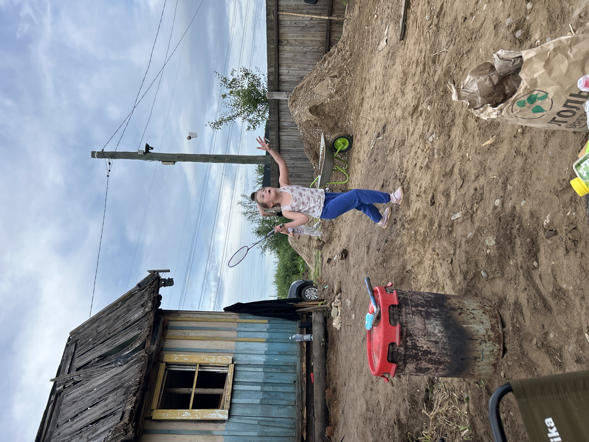 In Chaikovsky, Russia, a young girl, dressed in a white tank top and blue pants, is captured mid-motion as she plays badminton. She is positioned on a sandy, uneven ground with a dilapidated wooden structure to her left, characterized by a weathered roof and peeling paint. In the background, a wooden fence encloses a yard with piles of construction debris and a wheelbarrow. A utility pole with electrical wires stretches across the sky, which is overcast with clouds. The scene appears to be an outdoor area, possibly a backyard or a construction site, with various objects scattered around, including a red trash bin and a paper bag.