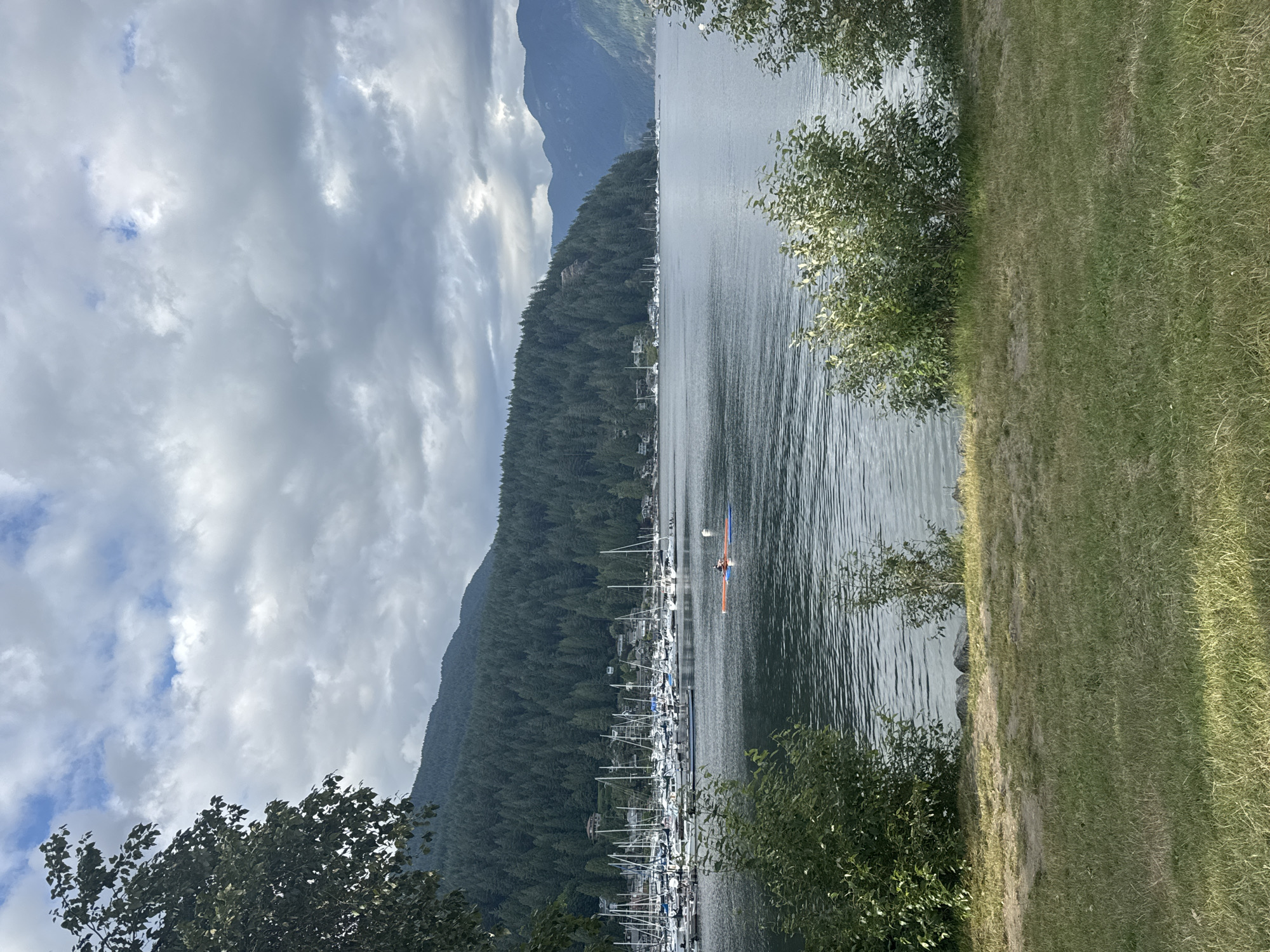 The image depicts a scenic view of a marina and coastline at Fisherman's Cove in the District of North Vancouver, Canada. In the foreground, a grassy area with some dirt patches leads down to the water's edge, with bushes framing the scene. The water itself is calm with ripples reflecting the cloudy sky. A marina filled with numerous sailboats is visible in the middle ground, lining the shore. Beyond the marina, a densely forested hillside rises, dotted with houses. In the distance, mountains form the backdrop, their peaks softened by the overcast weather. On the water in the middle ground, two people are visible on a small, inflatable raft, appearing to be paddling. The overall atmosphere is tranquil and natural, with a blend of human activity and the surrounding wilderness.