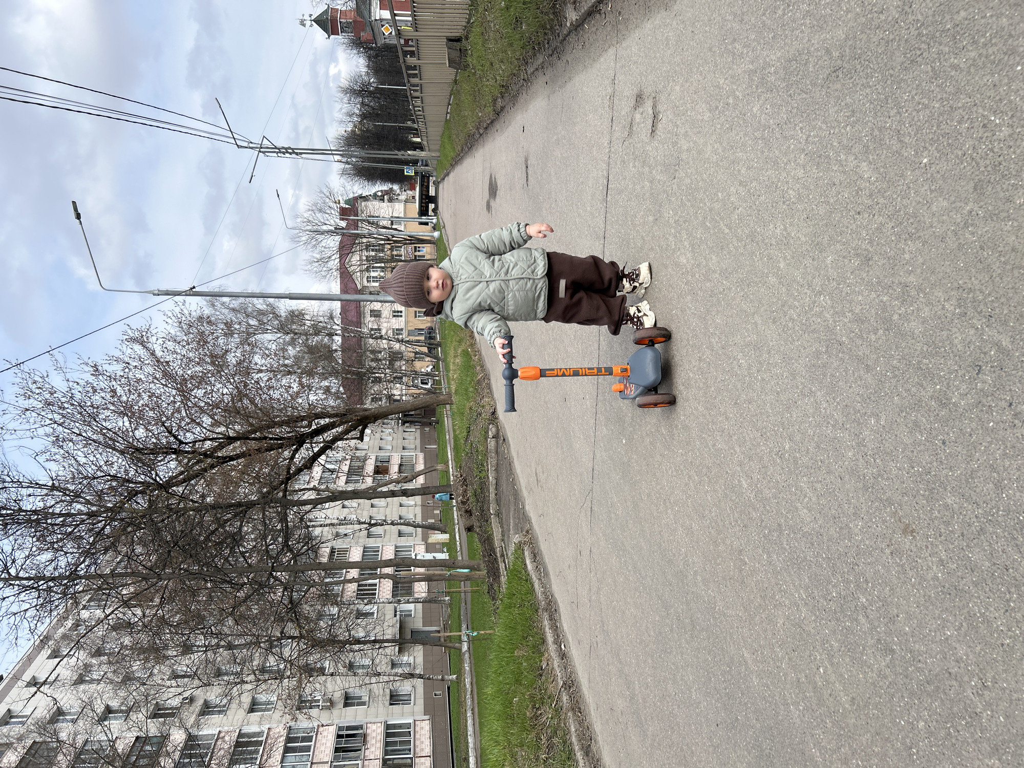 A small child, approximately two years old, stands on a gray paved path in Klin, Russia. The child wears a brown knit hat, a light green quilted jacket, and brown pants. The child is riding a three-wheeled scooter with an orange frame and gray wheels. The scooter has the word "TRIUMPH" printed on it. Buildings are visible in the background, including a tall apartment building with multiple rows of windows and a building with a red roof and a tower. Bare trees are on the left side of the image and the sky is overcast. Utility poles and wires stretch across the top of the image. Green grass is growing alongside the path.