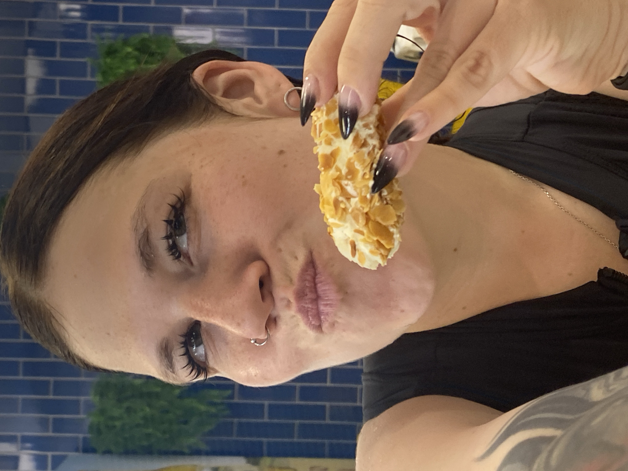 A medium close-up shot shows a woman with brown hair and light skin, with freckles on her nose and cheeks. She has long, dark eyelashes and is wearing a nose ring. Her mouth is pursed as if she is about to eat. She is holding a small, oval-shaped pastry covered in sliced almonds with her right hand. Her fingernails are long and painted in a gradient of black to clear. She is wearing a black tank top. The background is a blue tiled wall with green foliage visible to the left. The scene is indoors, and the lighting suggests it is daytime. This image was taken in Votkinsk, Russia.