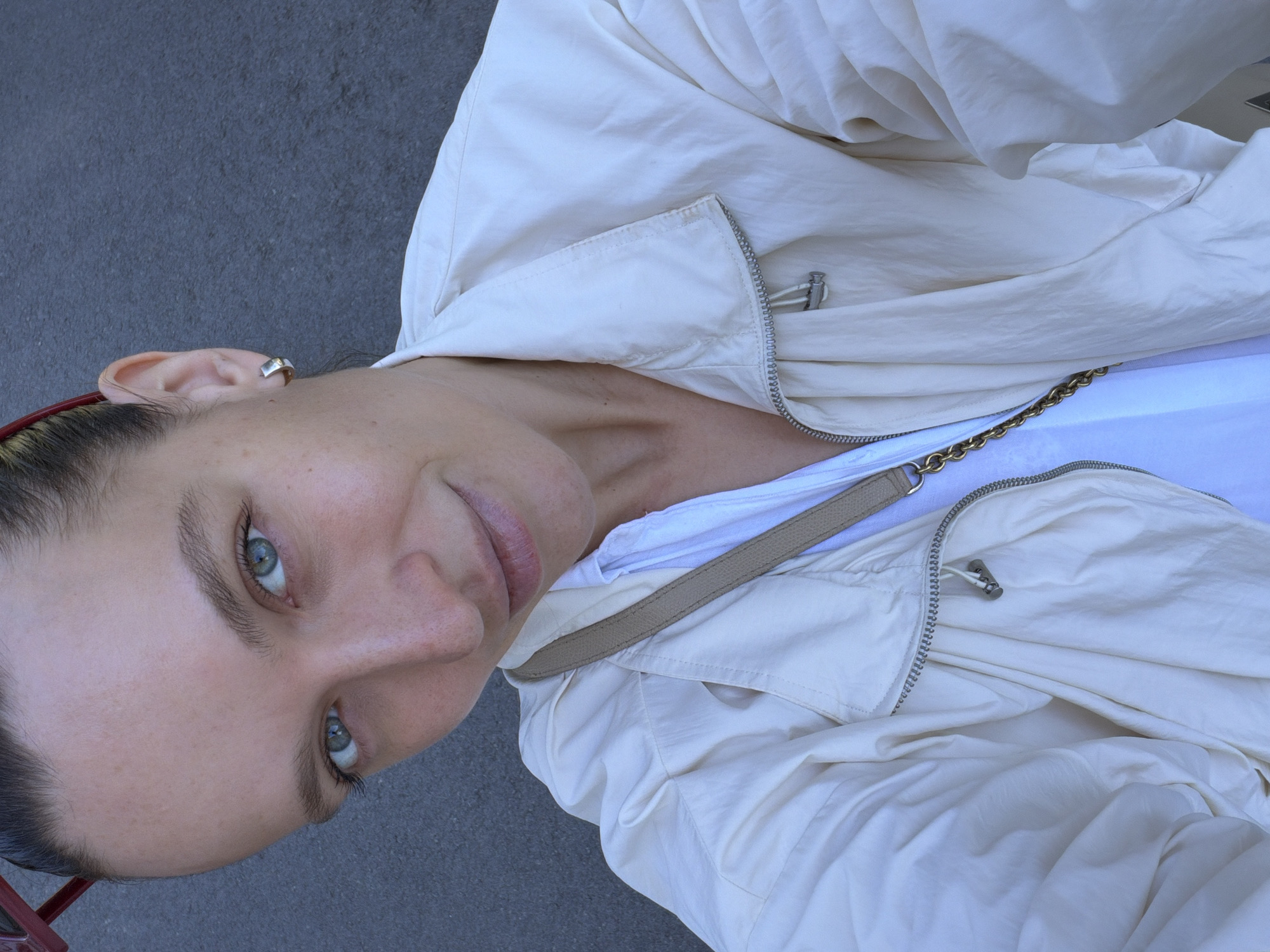 A selfie of a woman with fair skin, pulled-back dark hair, and light blue eyes, is oriented from a low angle, tilted to the left. She wears a white t-shirt and a partially unzipped, light beige jacket with a gold chain detail. A tan-colored bag strap crosses her chest. A silver earring is visible in her left ear. The background is a gray asphalt surface. The image was likely taken in Boreti, Montenegro, based on metadata.