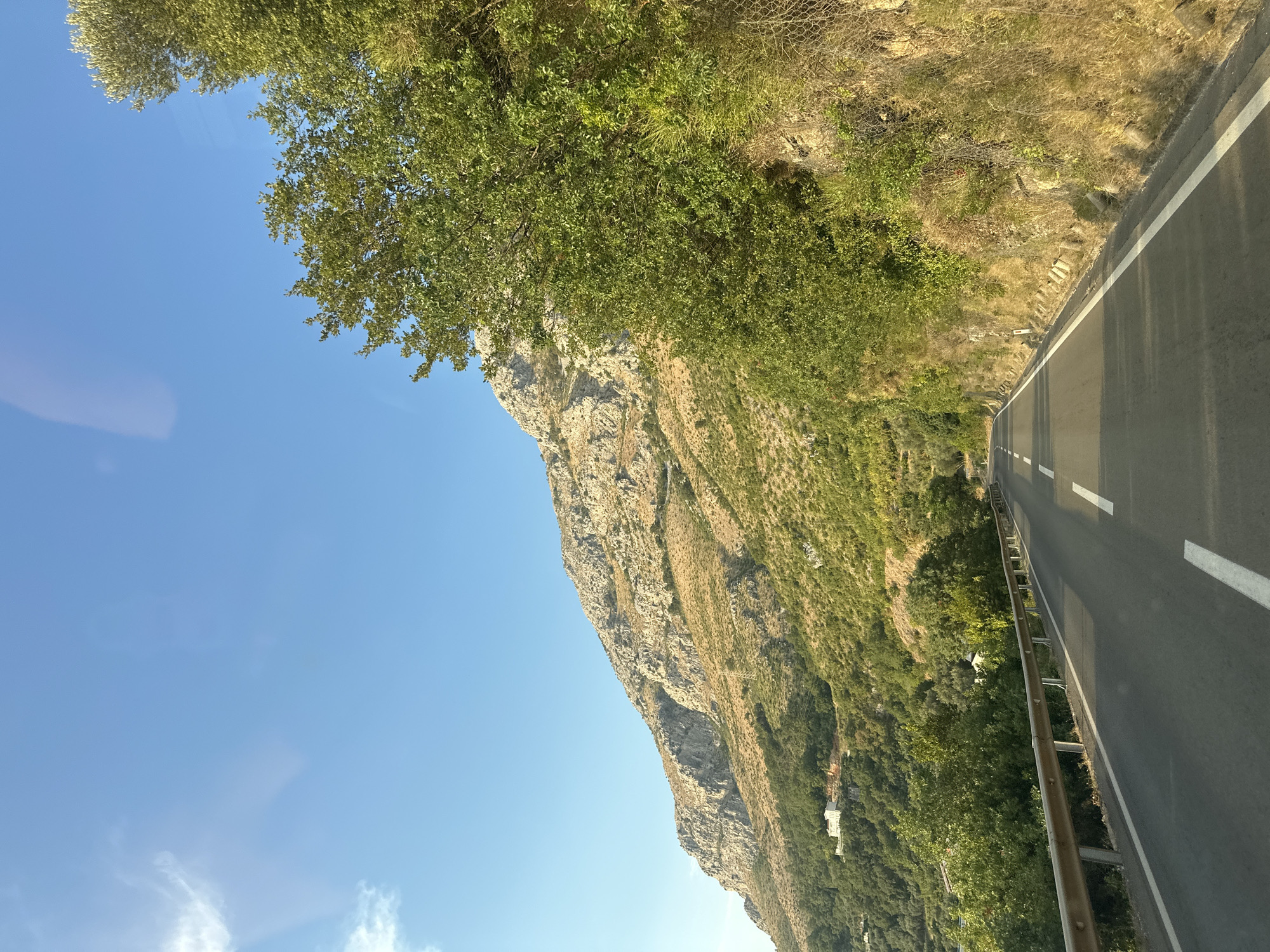 The image shows a winding road ascending a mountainous landscape in Rijeka Reževići, Montenegro. The road is paved and has white lane markings. A metal guardrail runs along the left side of the road. The mountainside is covered in lush green vegetation and rocky terrain. A few white buildings are visible nestled on the slopes. The sky is a clear blue with a few wispy clouds. The scene is captured from the perspective of someone traveling on the road, suggesting a journey through a scenic area. The overall impression is one of natural beauty and exploration.