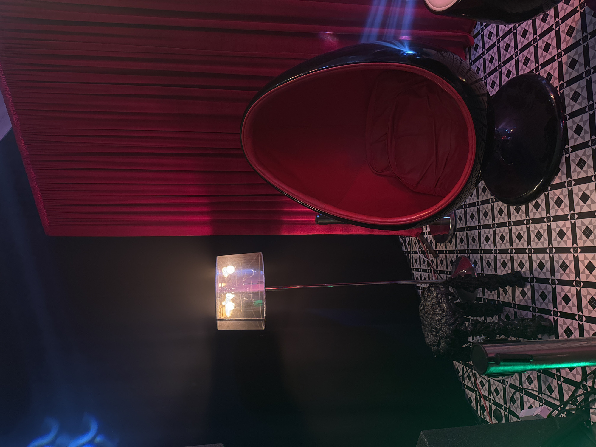 The image depicts an interior setting in Busmantsi, Bulgaria, featuring a predominantly black and red color scheme. A striking red egg-shaped chair with a black exterior stands prominently, positioned near a checkered black and white patterned wall. A floor lamp with a translucent shade illuminates a dark wall. A red curtain hangs in the background, adding to the dramatic ambiance. Some unusual textured object is visible near the checkered wall.