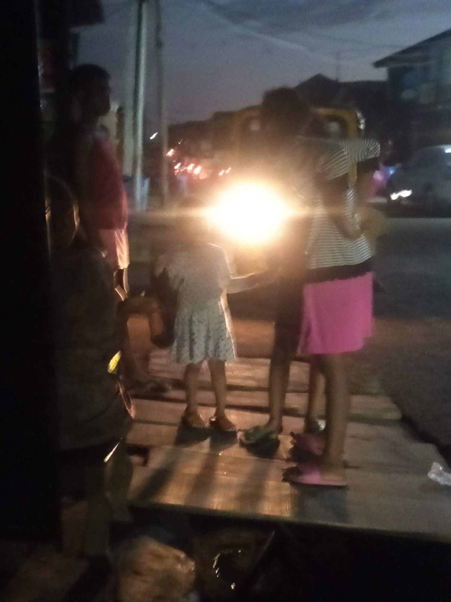 The image captures a nocturnal scene in Shangisha, Nigeria, featuring several individuals gathered on a wooden platform or walkway. A dominant, bright yellowish-white light source, likely from a vehicle's headlights or a strong street lamp, illuminates the center of the frame, causing significant lens flare and silhouetting many subjects.
On the far left, an adult or older child is partially visible. Next to them stands another individual wearing a red top and light-colored shorts, primarily seen as a silhouette. In the center-right, two younger children are present. One child wears a light-colored, possibly white, patterned dress, while another is dressed in a striped top and a vibrant pink skirt. Their figures are also partially obscured by the intense central light. Additional blurred figures and shapes suggest more people or objects further in the background.
The background itself is dark and out of focus, revealing blurred lights from what appear to be distant vehicles or streetlights, indicating an urban or semi-urban setting. The dark sky confirms the time of day as night or very late dusk. The wooden platform beneath the figures shows distinct planks, and various pieces of footwear, including flip-flops, are visible near their feet. No discernible text is present in the image. The scene depicts a casual gathering or simply people standing in an everyday setting under challenging low-light conditions.