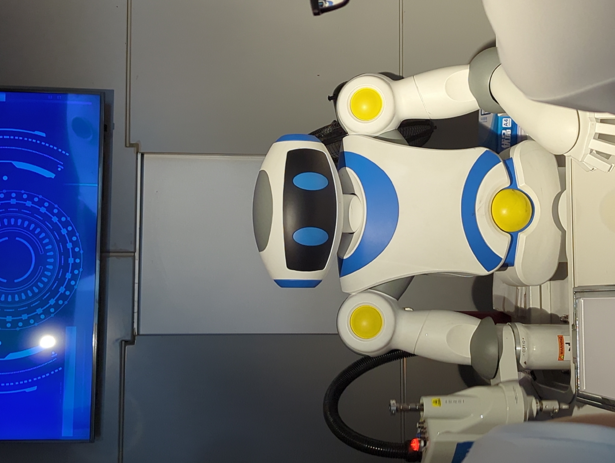 The image shows a white and blue robot with a gray head, standing in what appears to be a laboratory or exhibition space. The robot has a screen above it displaying a blue, circular graphic with technological elements. To the left of the robot, there is a white robotic arm with a black hose attached. In the foreground, part of a person's shoulder and arm are visible, wearing a light blue shirt. The robot is positioned in front of a gray wall and what looks like a white counter or table. There is also a stack of what appears to be paper, one of which has "A4" printed on it. A warning sticker with a red symbol is visible on a cylindrical object near the base of the robot. The overall impression is of a modern, technologically advanced setting, possibly related to robotics or automation. There is no specific indication of the location being 江北城街道, China, other than the prompt's suggestion.