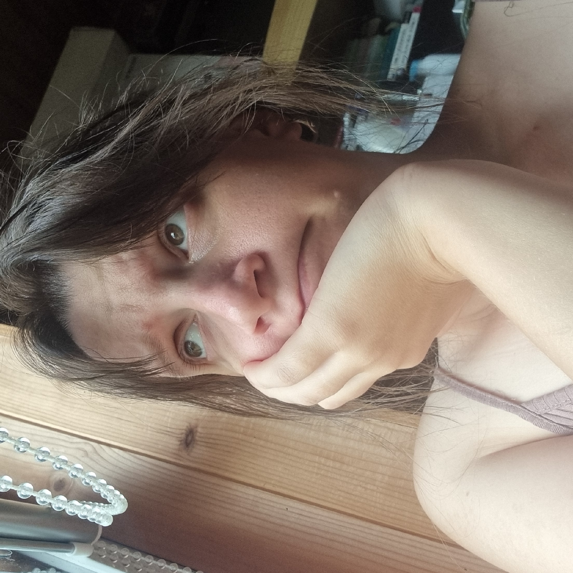 A close-up of a woman's face shows her looking upwards with a bored expression, her right hand propping up her chin. She has brown hair and is wearing a light brown tank top. The background is a wooden wall, with beaded curtains to the left and shelves of objects to the right. The image is taken indoors, likely in a home environment. This scene does not provide any specific information about the location being Dunnae-myeon, South Korea.
