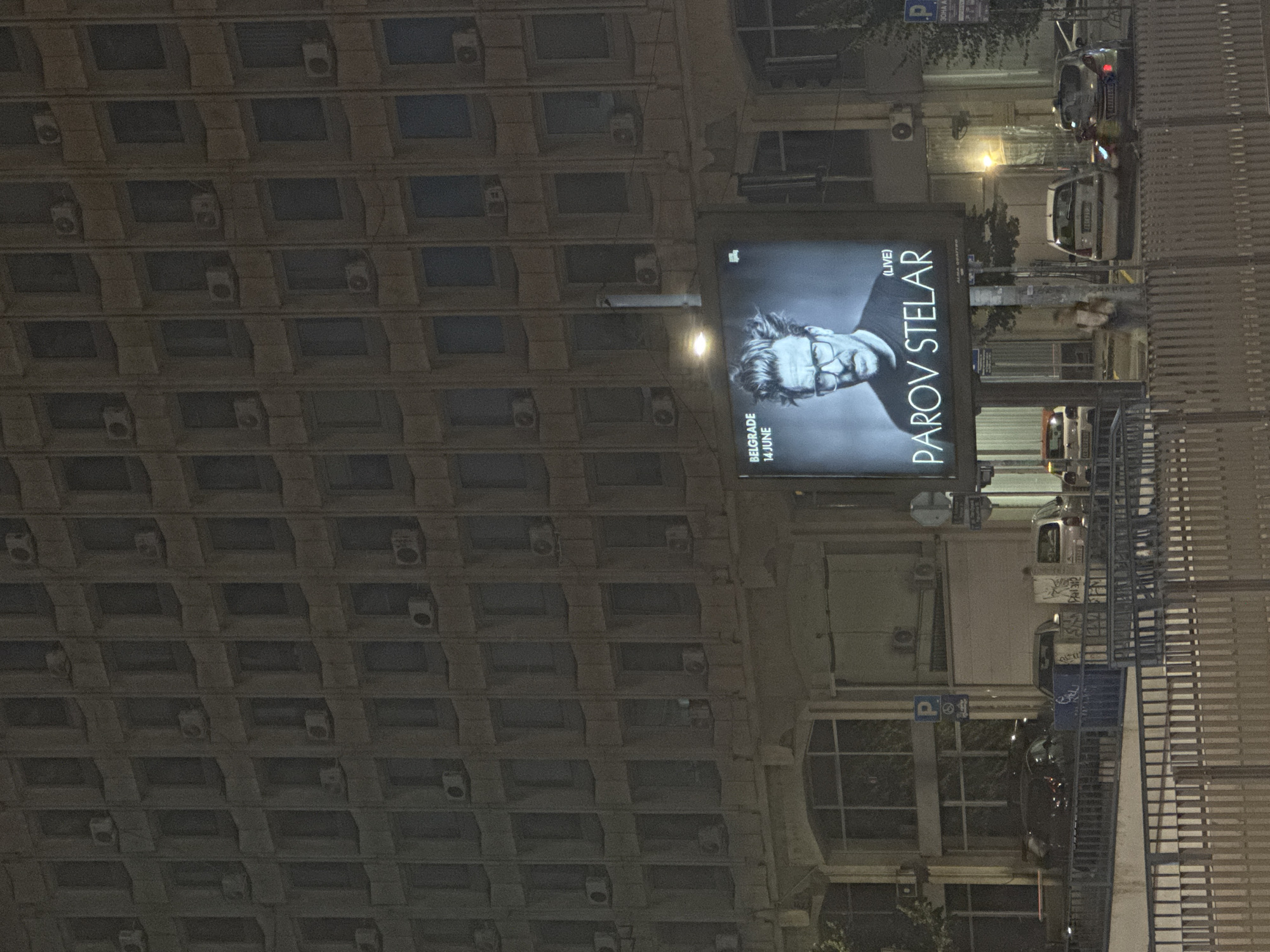 An overhead, slightly angled nighttime photograph captures a large, multi-story building and an illuminated digital billboard in Belgrade, Serbia. The prominent building features a grid-like facade composed of numerous recessed rectangular windows, with a light brown or grey concrete exterior. Many white, wall-mounted air conditioning units are affixed below the windows on various levels. The ground floor of the building has larger, arched window sections.
In the mid-ground, a brightly lit digital billboard displays a black and white portrait of a man with glasses and disheveled hair. Text on the billboard reads "BELGRADE 14 JUNE", "(LIVE)", and "PAROV STELAR".
The immediate foreground is occupied by a dark-colored metal fence with vertical slats. Behind this fence and the billboard structure, a street scene is visible. Several cars are parked or moving; these include a white hatchback (potentially a Renault Clio or similar) with a partial Serbian license plate visible on the right, and a silver hatchback (possibly a Fiat Punto) with a partially visible Serbian license plate "BG 144..." on the far right. A dark-colored vehicle is parked on the left. A single person, possibly a woman, walks behind the billboard structure, illuminated by its light. Blue street signs, including a "P" for parking with an accessibility symbol, are visible on the right side of the road. The overall environment is dimly lit by ambient street lighting and the billboard itself.