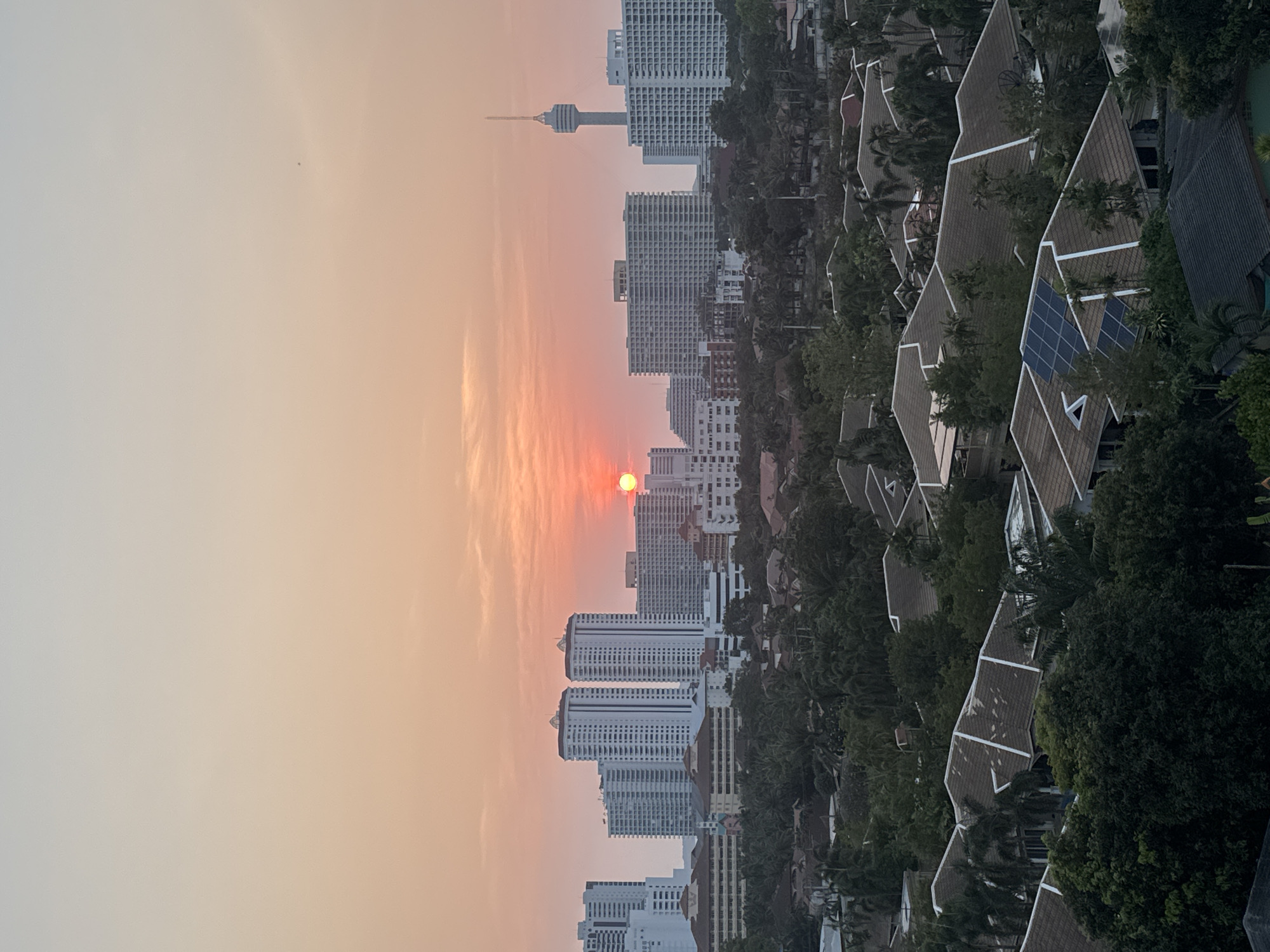 The image shows a sunset over Pattaya City, Thailand. The sun is a red-orange orb positioned in the sky above a line of high-rise buildings. These buildings are primarily white and rectangular. In the distance is a tall tower. In the foreground, there are angled rooftops of houses with solar panels installed on some of them. There are also trees interspersed with the buildings. The sky is a gradient from light grey to orange closer to the sun.