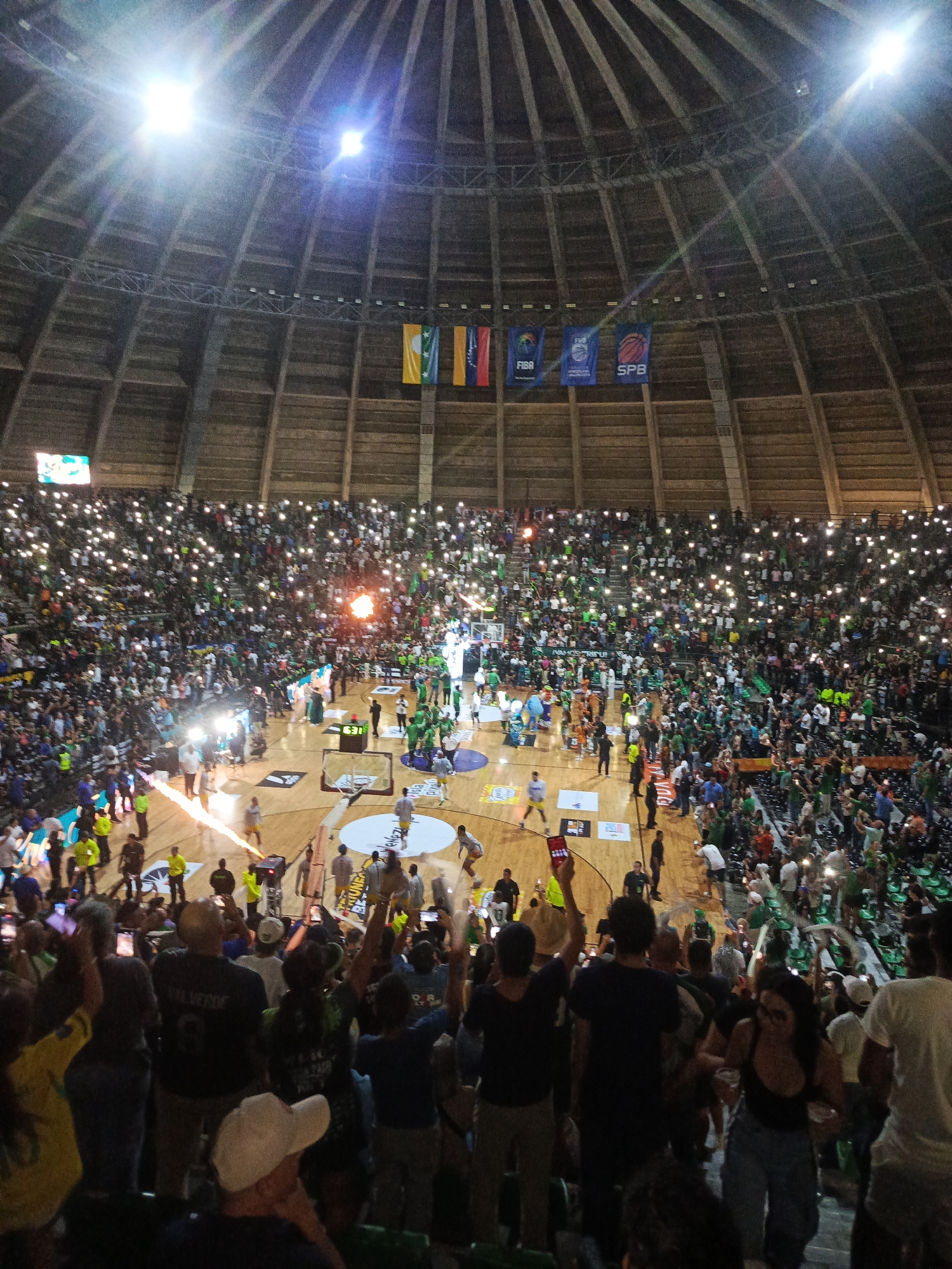 The image depicts a packed basketball arena during an event. The dome-shaped ceiling is visible, supported by wooden beams, with bright lights illuminating the space. Numerous flags hang from the upper structure. The stands are filled with a large crowd, many holding up their phones with lights on, creating a sea of small lights throughout the arena. The basketball court below is partially visible, with players and staff on it. There are also pyrotechnic displays or flames being emitted from devices on the court, adding to the dramatic atmosphere. The scene appears to be a lively and enthusiastic gathering, likely a sporting event or concert. The mention of the city of La Asuncion, Venezuela, suggests this is the location of the depicted event, though there are no overt visual cues within the image to confirm this specific location.