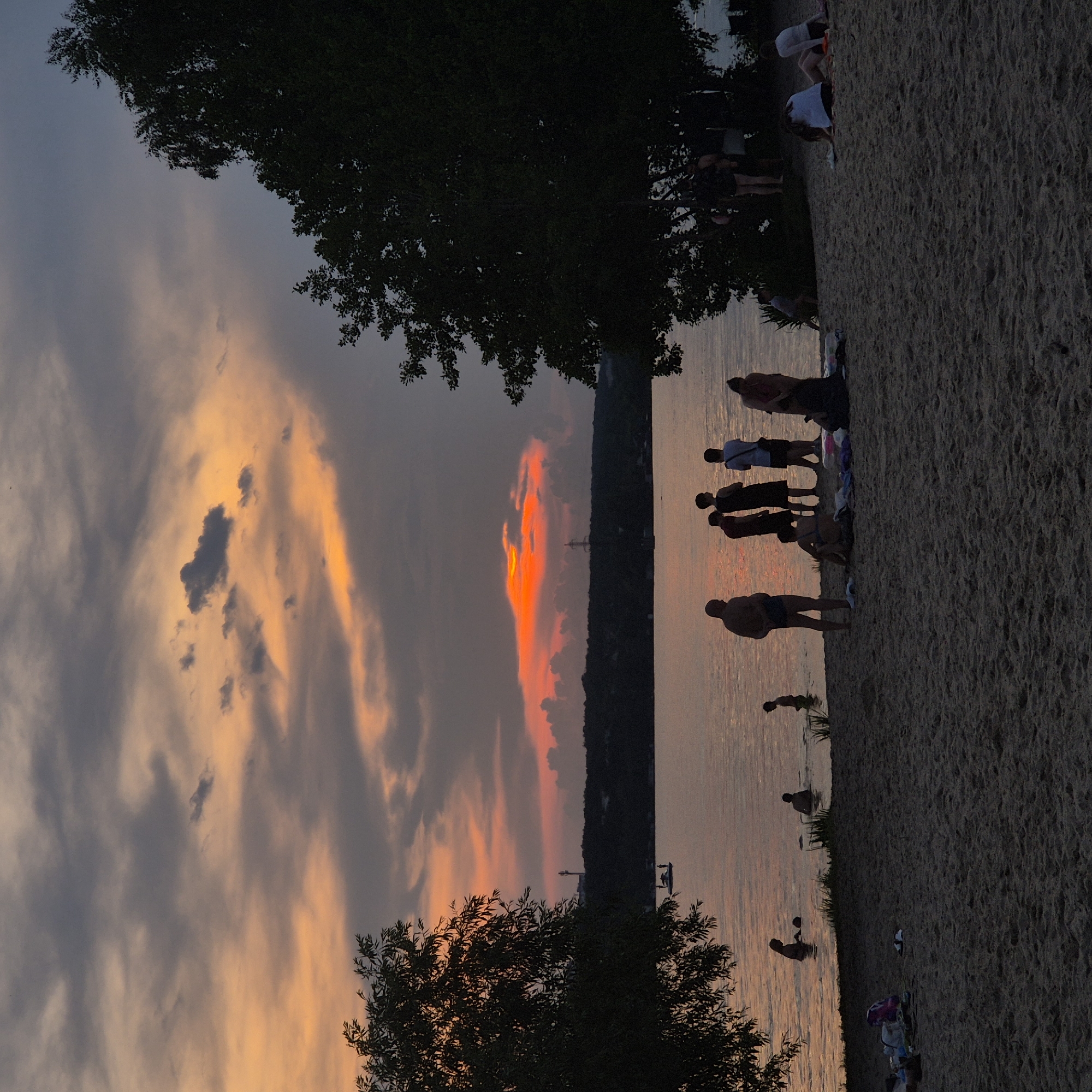 The image depicts a sunset scene at a beach located on the Voronezh Reservoir in Voronezh, Russia. Silhouettes of people are visible on the sandy shore and in the water. Some are standing, others are sitting or wading. A group of individuals stands on the beach facing the water, while others are in the shallow part of the reservoir. The sky is filled with dramatic clouds, illuminated by the warm hues of the setting sun, with vibrant orange and pink tones contrasting with darker grays. Trees frame the scene on the left and right, casting shadows. A small boat with figures on it is visible on the water in the distance, as is a silhouette of a distant shoreline with buildings and a tower. The atmosphere is serene, capturing the end of a day at the reservoir.