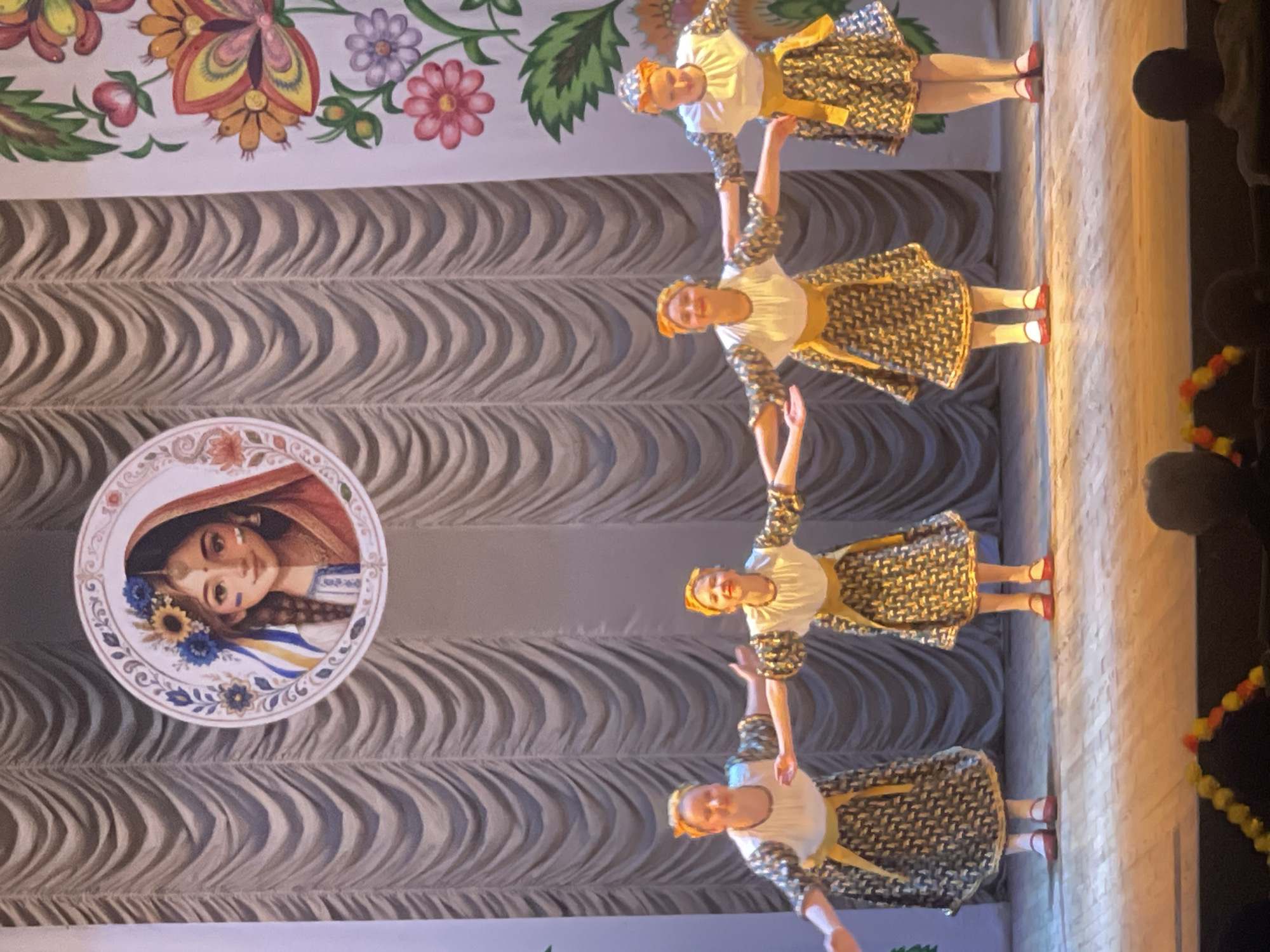 In Horbanivka, Ukraine, four young girls, dressed in traditional Ukrainian folk costumes, are performing on a stage. The costumes consist of white blouses with puffed sleeves, black and gold patterned skirts, and yellow sashes. They are also wearing orange headbands. The girls stand in a line, facing the audience, with their arms extended outwards in a synchronized dance pose. The stage is adorned with a backdrop featuring grey curtains and a circular image of a woman with Ukrainian flag colors incorporated into her hair. To the right of the stage, a colorful floral painting adds to the festive atmosphere. The lighting on stage is bright, illuminating the performers. In the foreground, the audience is dimly visible, seated in a theater.