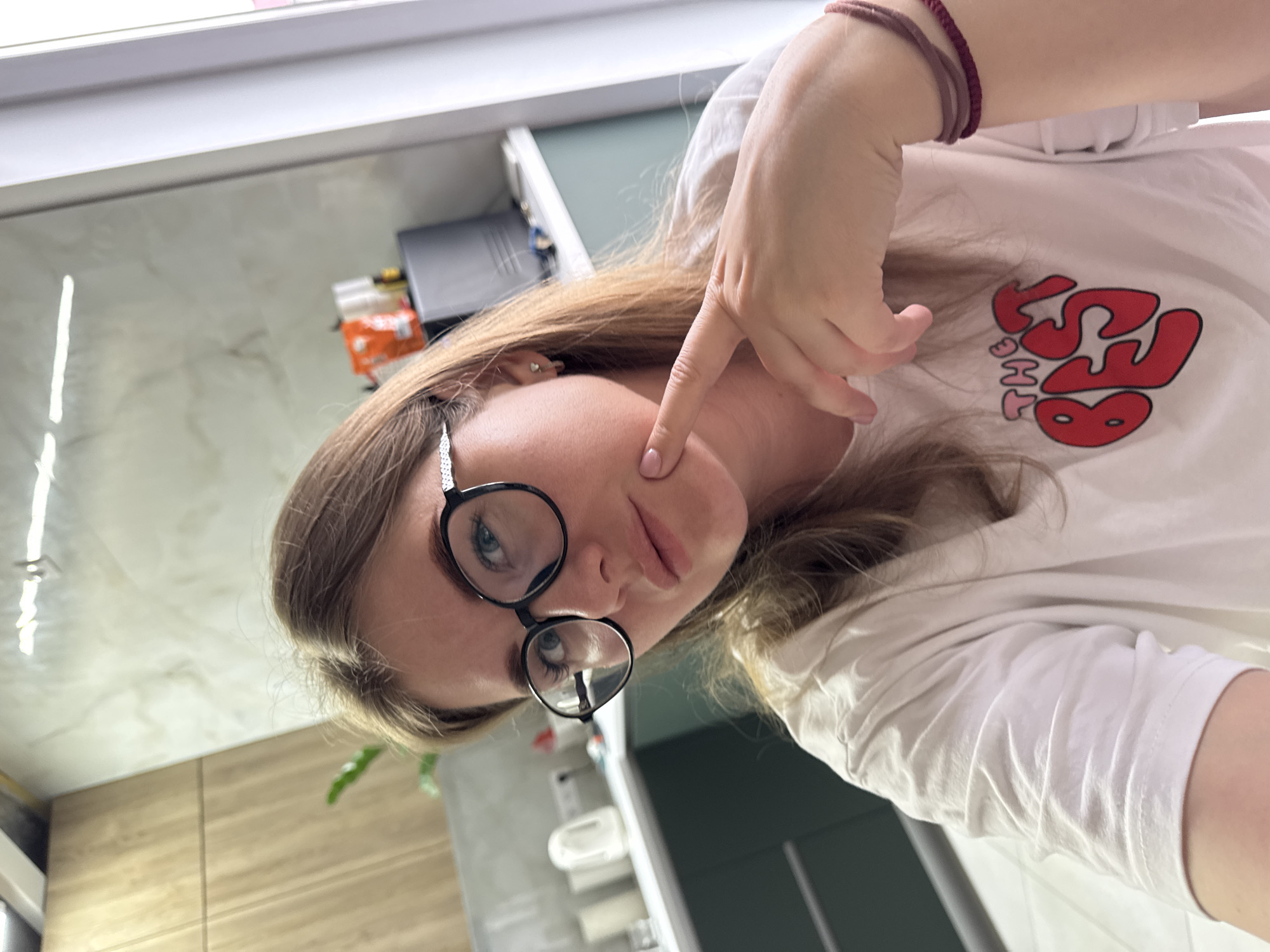 A selfie of a woman with light brown hair wearing black-rimmed glasses and a white t-shirt with a red and pink logo. Her finger is touching her cheek. The background includes a countertop, cabinets, and a partially visible toilet. The specific location in Gornyi (Dvurechenskoe S/P), Russia cannot be determined from this image alone.