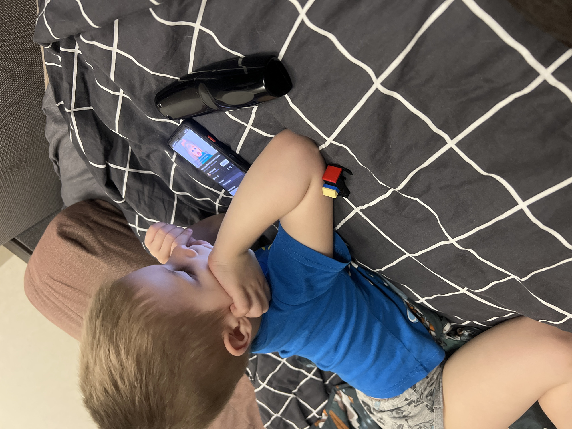 A young child is lying on a bed or sofa in Chaikovsky, Russia, engrossed in a smartphone. The child, a boy with light brown hair, is wearing a blue t-shirt and grey patterned shorts. He is lying on his side, with his right hand near his face, possibly resting or indicating concentration. The bed is covered with a black duvet featuring a white grid pattern.
Next to the child on the blanket is a black smartphone, its screen displaying a video, likely from a streaming platform, with a cartoon-like character visible. Visible Russian text on the screen includes "Обычные видео" (Normal videos) and buttons like "Нравится", "Дизлайк", "Поделиться" (Like, Dislike, Share), suggesting YouTube. The time "08:00" is displayed in the top left corner of the phone. Near the phone, there is a glossy black, cylindrical object, possibly a remote control or a portable speaker. Also on the blanket, close to the child's arm, is a small toy composed of red, blue, and yellow building blocks, resembling Lego. The lighting suggests an indoor setting, possibly during the evening.