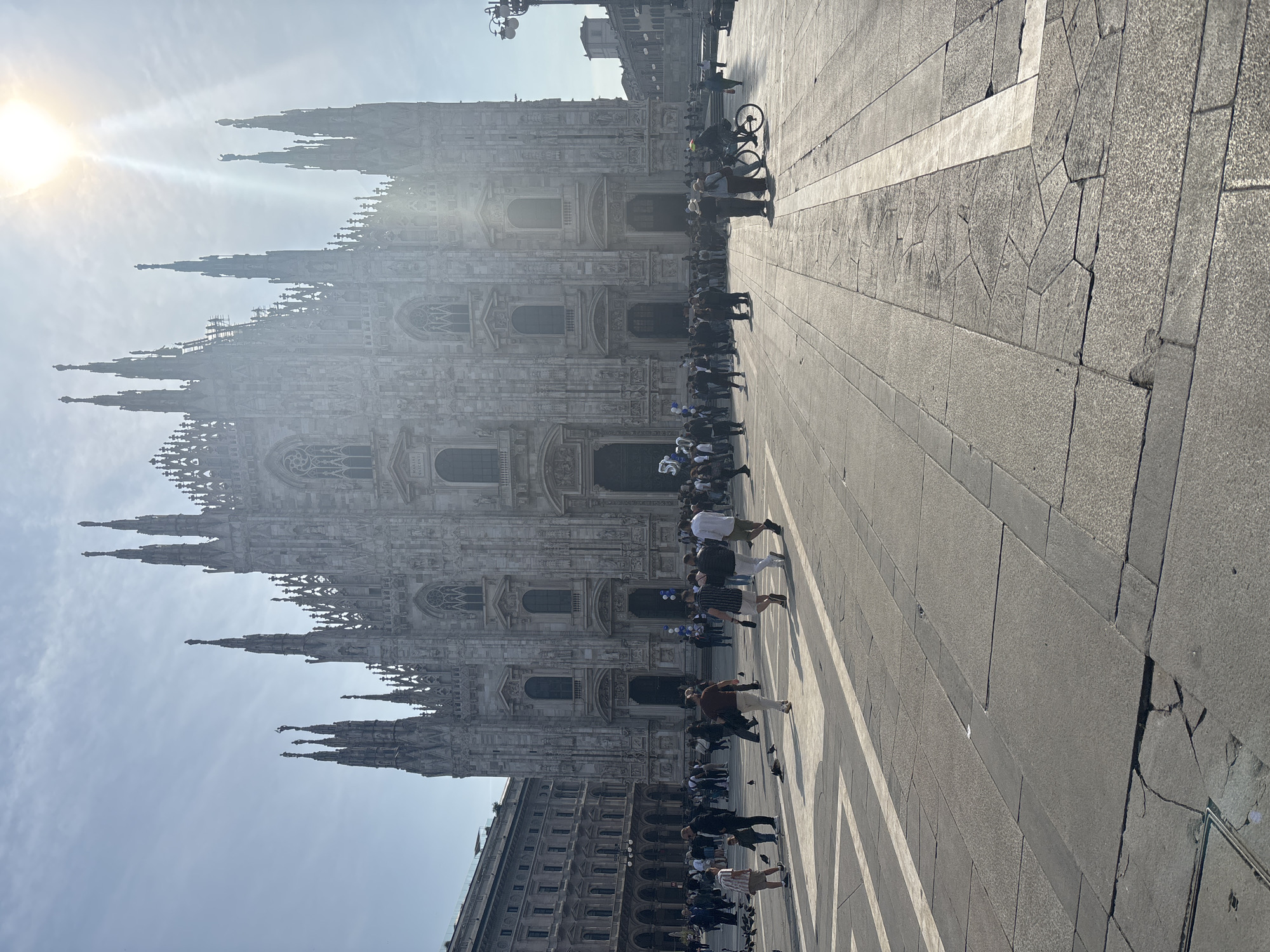 The image captures the Milan Cathedral (Duomo di Milano) in Milan, Italy. The cathedral's facade is prominently displayed, with its spires reaching towards the sky. A group of people is present in the foreground, walking along the paved plaza in front of the cathedral. Some individuals are walking alone, while others are in small groups. A bicycle is also visible. The environment is bright, with the sun shining in the sky.