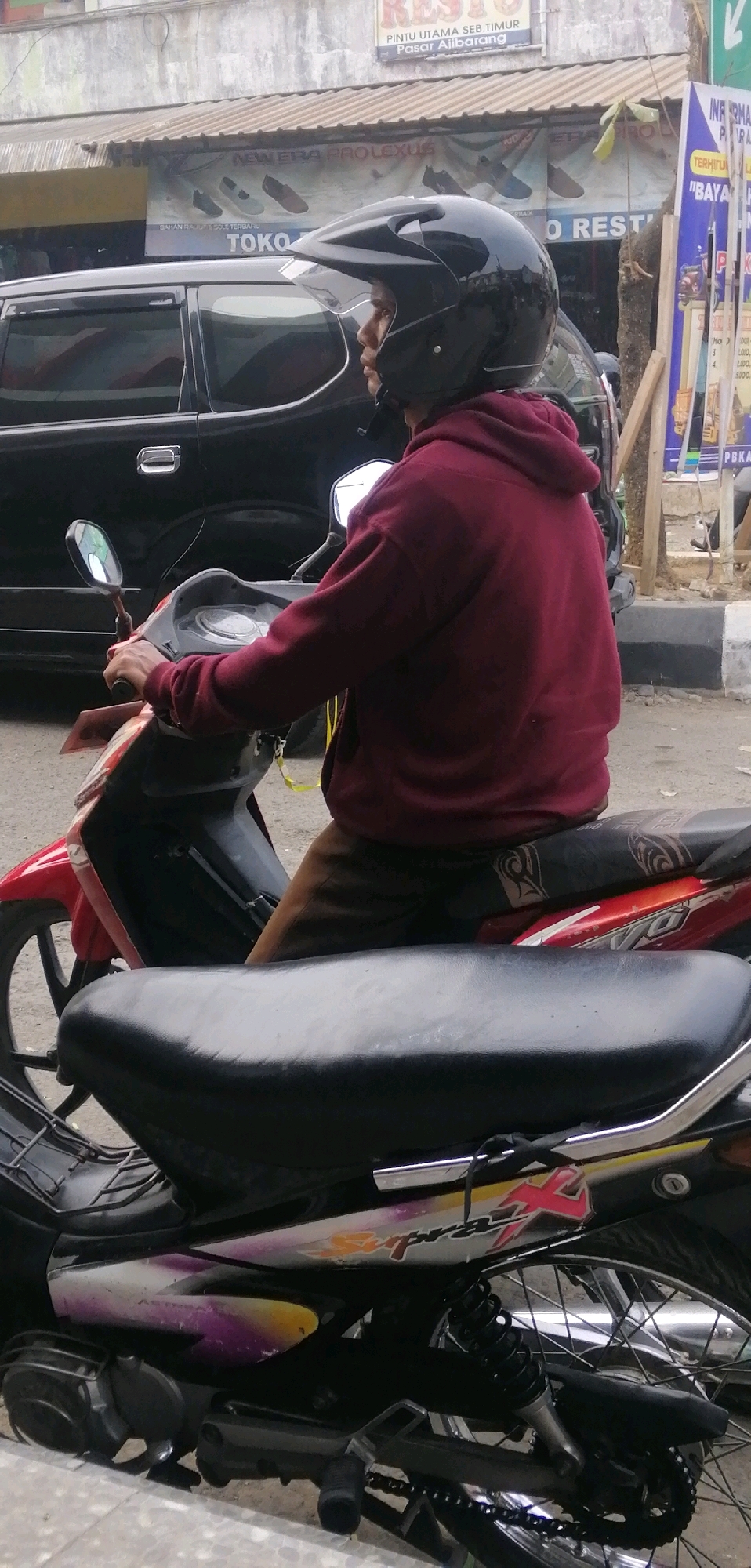 The image shows a person wearing a black helmet and a maroon hoodie on a red and black motorcycle. The person is seated on the motorcycle, holding the handlebars, and looking to the right. The motorcycle is parked on a street in Ajibarang, Indonesia, as indicated by a sign in the background that reads "Pintu Utama Seb. Timur Pasar Alibarang". Behind the motorcycle, a black car is visible. There are also storefronts with various signs in the background, including one advertising "NEW ERA PROLEXUS" and another with text in Indonesian that appears to be information or advertisements. The environment appears to be an urban or semi-urban street scene with some greenery visible. The time of day appears to be daytime, with moderate lighting.