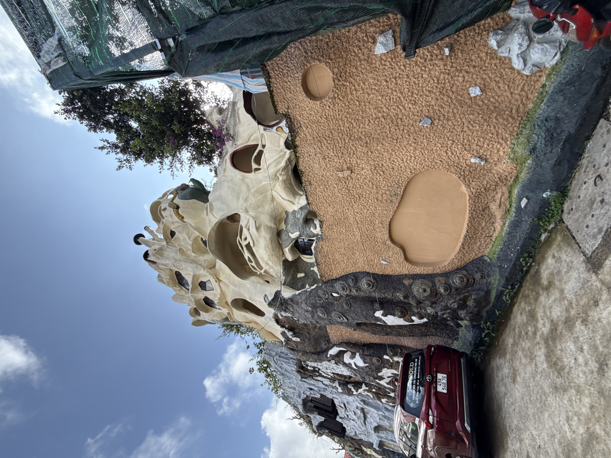 The image, oriented upright, showcases the distinctive and organic architecture of the "Crazy House" (Hằng Nga Guesthouse) in Da Lat, Vietnam, under a bright, sunny sky. The multi-story building features a whimsical, sculpted design resembling giant tree trunks, caves, and natural formations. One prominent section is a light, cream-colored structure with numerous irregular windows and openings, giving it a cave-like appearance. Adjacent to this, darker, rugged surfaces mimic tree bark or rock, with swirling patterns and textural variations. A large section of the building's exterior is covered in a reddish-brown, granular texture, featuring circular indentations.
Below the unusual architecture, a red car, identifiable as a Toyota MPV (likely an Innova), is parked on a concrete ground. The car's rear displays the license plate "49A 548.76". A white sticker on the back window contains the text "SALON GOLD" followed by Vietnamese text: "CHUYÊN NỐI TÓC CÔNG NGHỆ MỚI, VIP" (Specializing in new technology hair extensions, VIP) and a partially visible phone number "0979.xxxx.xxx".
The scene is set outdoors during the daytime, with a clear blue sky and scattered white clouds. Green trees and foliage are visible around and behind parts of the building, enhancing its natural, fantastical aesthetic. Sections of the view are partially obscured by dark green mesh netting, possibly used for shade or construction. The overall atmosphere is one of a unique and imaginative landmark within a sunny environment.