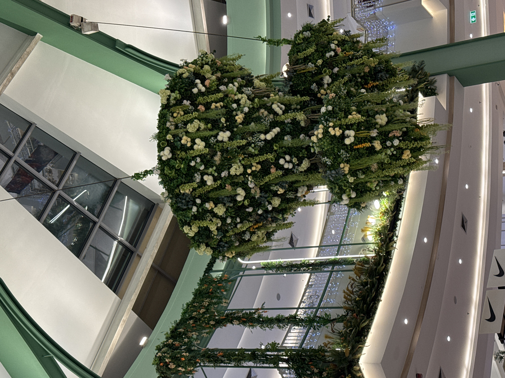 The image depicts a large, multi-tiered floral installation suspended from the ceiling of an indoor space, likely a shopping mall or atrium, in Patong, Thailand. The installation is predominantly composed of green foliage and various white, cream, and pale yellow artificial flowers and hanging greenery. The structure is lush and voluminous, with cascading elements creating a dramatic effect.
The surrounding environment features architectural elements consistent with modern retail spaces. A sloped ceiling with skylight panels is visible at the top, reflecting light. Large, pale green structural beams and columns frame the scene, adding to the decorative aesthetic. Below the floral installation, there appears to be a multi-level interior with balconies or walkways, lined with decorative lights. In the lower part of the image, sections of the ceiling and suspended banners with Nike logos are visible, confirming the location as a commercial establishment. The overall impression is one of opulent, nature-inspired interior decoration within a commercial setting. There are no people visible in the image.