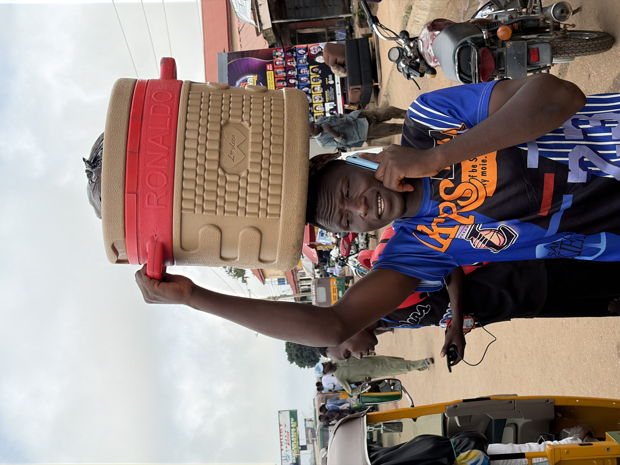 The image depicts a street scene in Gura Top, Nigeria, with a man carrying a large, tan and red cooler labeled "RONALDO" on his head. He is on his phone, and appears to be in conversation. He is wearing a blue and black t-shirt with a basketball-themed design and the number "23". In the foreground, a yellow and grey motorized rickshaw is visible. Behind the man, other people and motorcycles are present, and a large poster featuring portraits of many individuals is displayed. The environment appears to be a busy urban street with shops and stalls lining the road. The sky is overcast.