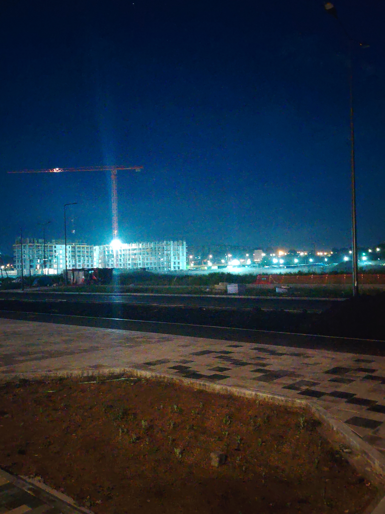 The image depicts a construction site at night in Yekaterinburg, Russia. A large tower crane, illuminated by lights, is visible in the center-left of the frame, its boom extending horizontally across the dark blue sky. Below the crane, a multi-story building under construction is brightly lit, with visible structural elements and numerous windows. Several smaller structures, possibly temporary site buildings, are situated at the base of the main building. In the background, a line of distant lights suggests other urban development or a city skyline. In the foreground, a paved area transitions into a patch of dark soil with sparse vegetation, bordered by a paved walkway with a patterned surface. A tall lamppost stands on the right side of the image. The overall scene conveys a sense of ongoing construction activity in an urban environment during nighttime hours.
