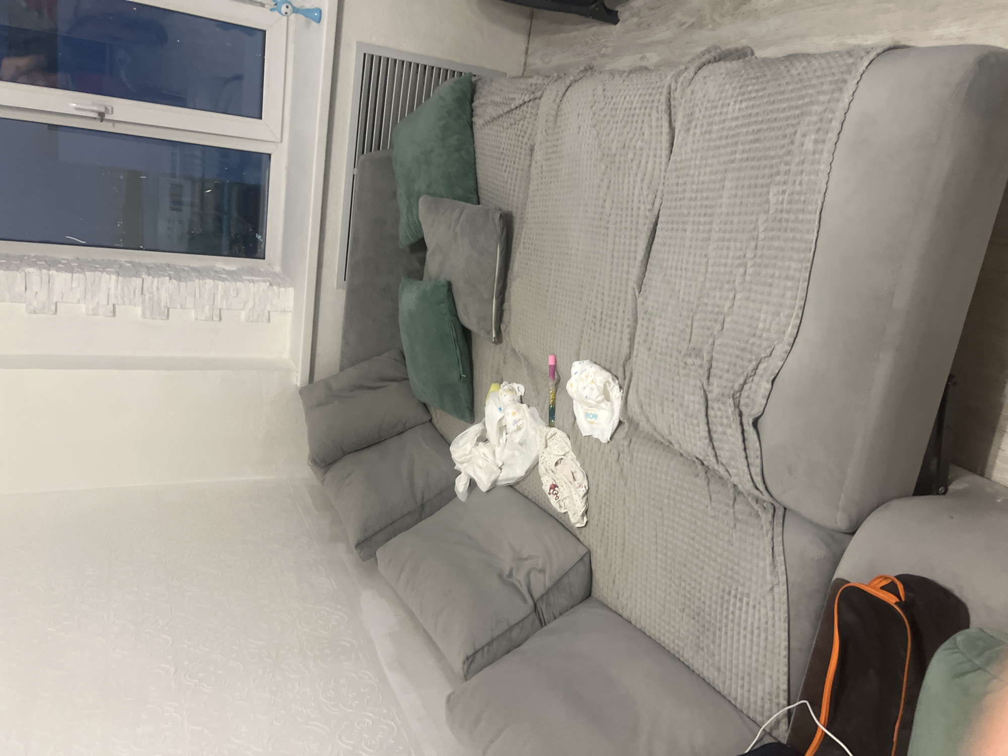 The image depicts an interior scene of a living room with a large grey sectional sofa. The sofa is covered with a grey waffle-weave blanket and several cushions, including two dark grey square ones and two emerald green ones. Scattered on the sofa are several baby items: a pile of white tissues, a small patterned garment that appears to be baby clothing, a diaper, and two colored markers. In the background, a window with a white frame is visible, reflecting the outside environment which appears to be a residential area. Next to the sofa, a black bag with orange trim is partially visible. The walls are white, with one wall featuring textured wallpaper with a subtle pattern. A white air vent is located below the window. The floor is a light wood laminate. The overall impression is a lived-in domestic space, possibly a nursery or a child's play area, given the presence of baby items. The scene is well-lit, likely by artificial light from inside the room and natural light from the window. The image does not provide any specific geographic indicators that would pinpoint its location to Veteran-2, Russia, or any other specific city.