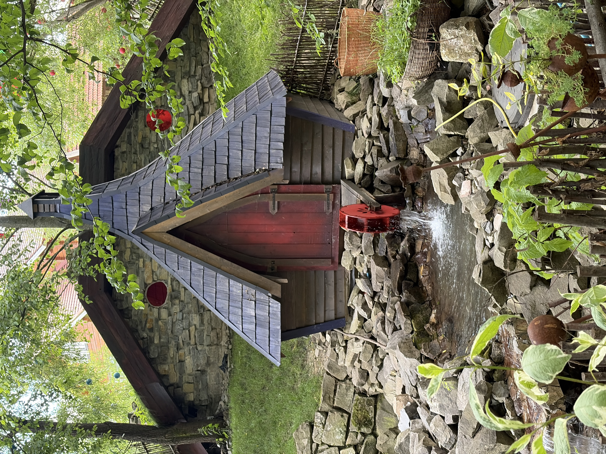This is a photograph of a rustic garden feature that includes a small, wooden structure with a red door, possibly a playhouse or shed, integrated into a stone wall. The structure has a dark, shingled roof. A red water wheel is positioned next to the structure, with water cascading over rocks, creating a small waterfall effect. Lush green foliage from trees and plants surrounds the scene, with some branches hanging into the frame. Red decorative orbs are visible hanging from the branches, and a woven basket is placed on top of the stone wall. The overall impression is one of a charming, possibly whimsical, garden. The lighting suggests it is daytime, and the presence of water and vibrant greenery indicates a pleasant, possibly humid, weather condition. There is no visible text in the image.