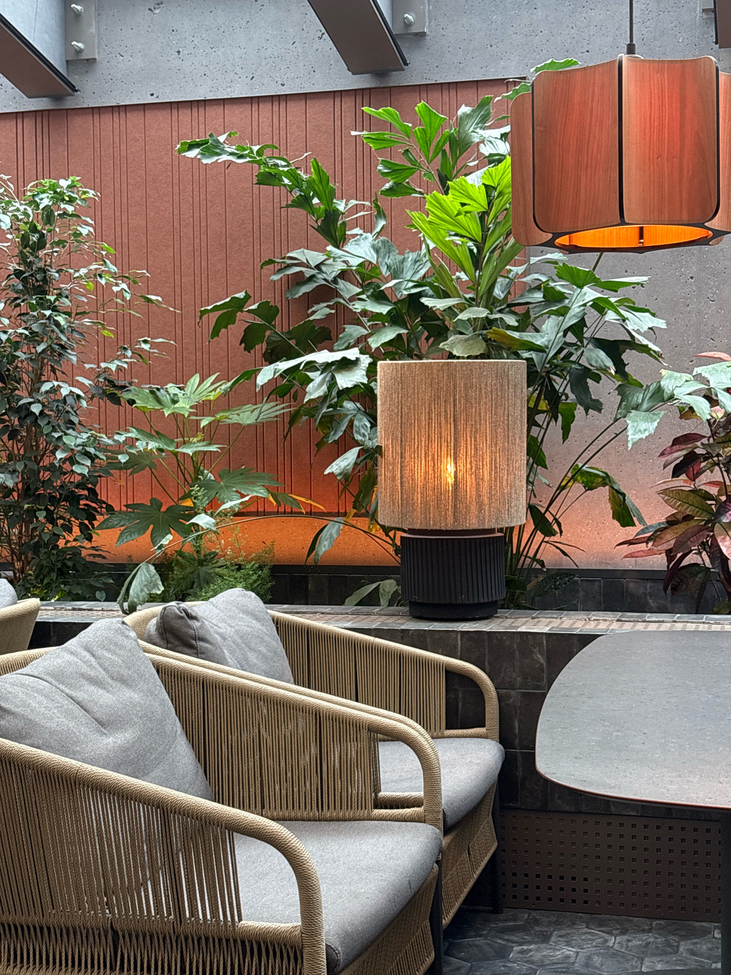 This is an indoor scene with a lush collection of plants. In the foreground, two chairs with light gray cushions are visible, constructed from woven rope in a natural, tan color. Adjacent to the chairs is a dark, oval-shaped tabletop. Behind the chairs and plants, a wall with vertical brown paneling serves as a backdrop. A cylindrical table lamp with a textured, beige shade stands on a dark base, emitting a warm light, positioned amongst the greenery. Further back, a modern wooden pendant lamp hangs from the ceiling. The floor is covered with hexagonal tiles. The overall environment suggests a stylish, possibly restaurant or lounge area. The location is identified as Kopavogur, Iceland.