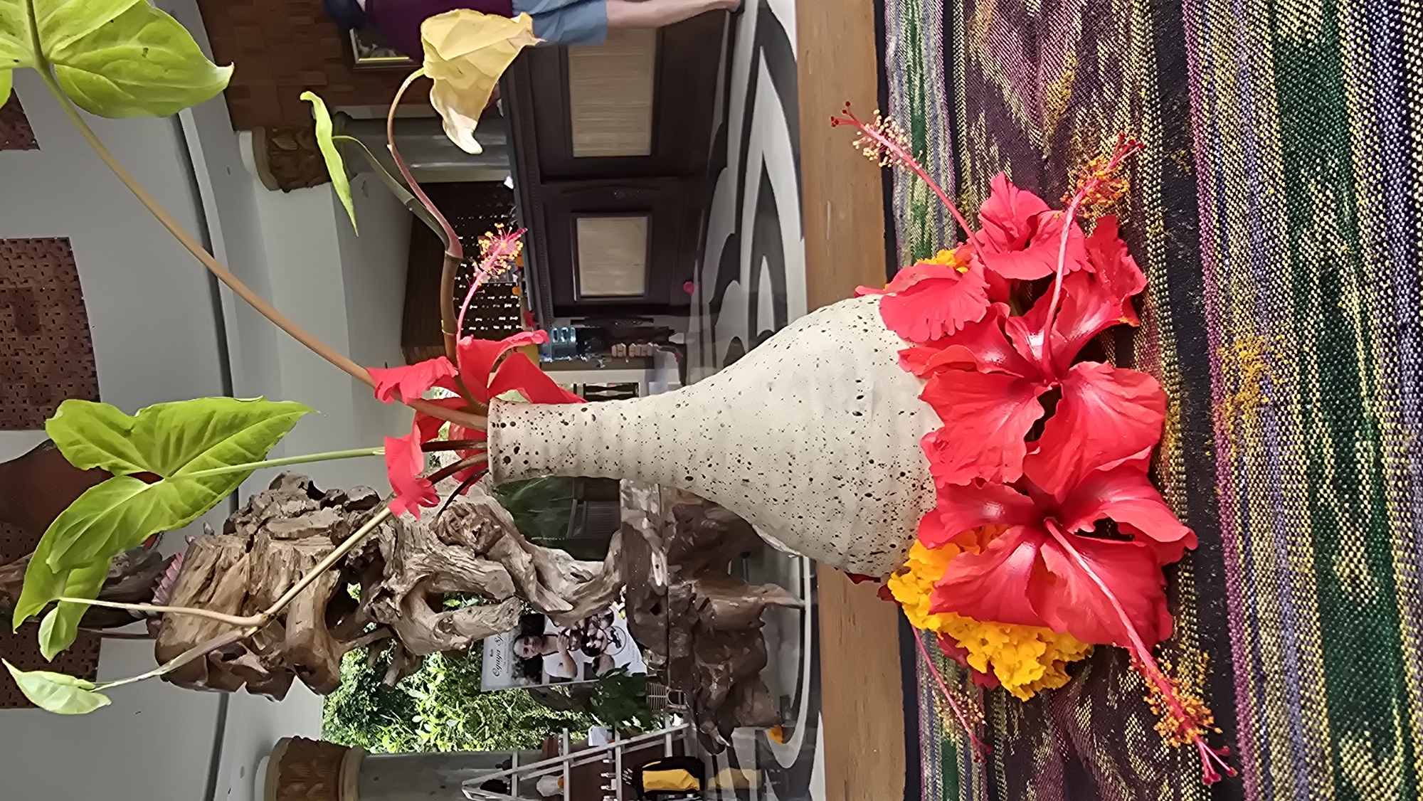 The image shows a floral arrangement likely located in the Ubud District, Indonesia. The arrangement features a textured, speckled vase filled with red hibiscus flowers, yellow marigolds, and leafy green plants, with a piece of driftwood positioned near the base. The background includes a striped fabric or textile, and reflections of interior features suggest a location indoors, possibly a hotel, gallery, or home in Ubud, known for its arts and culture.