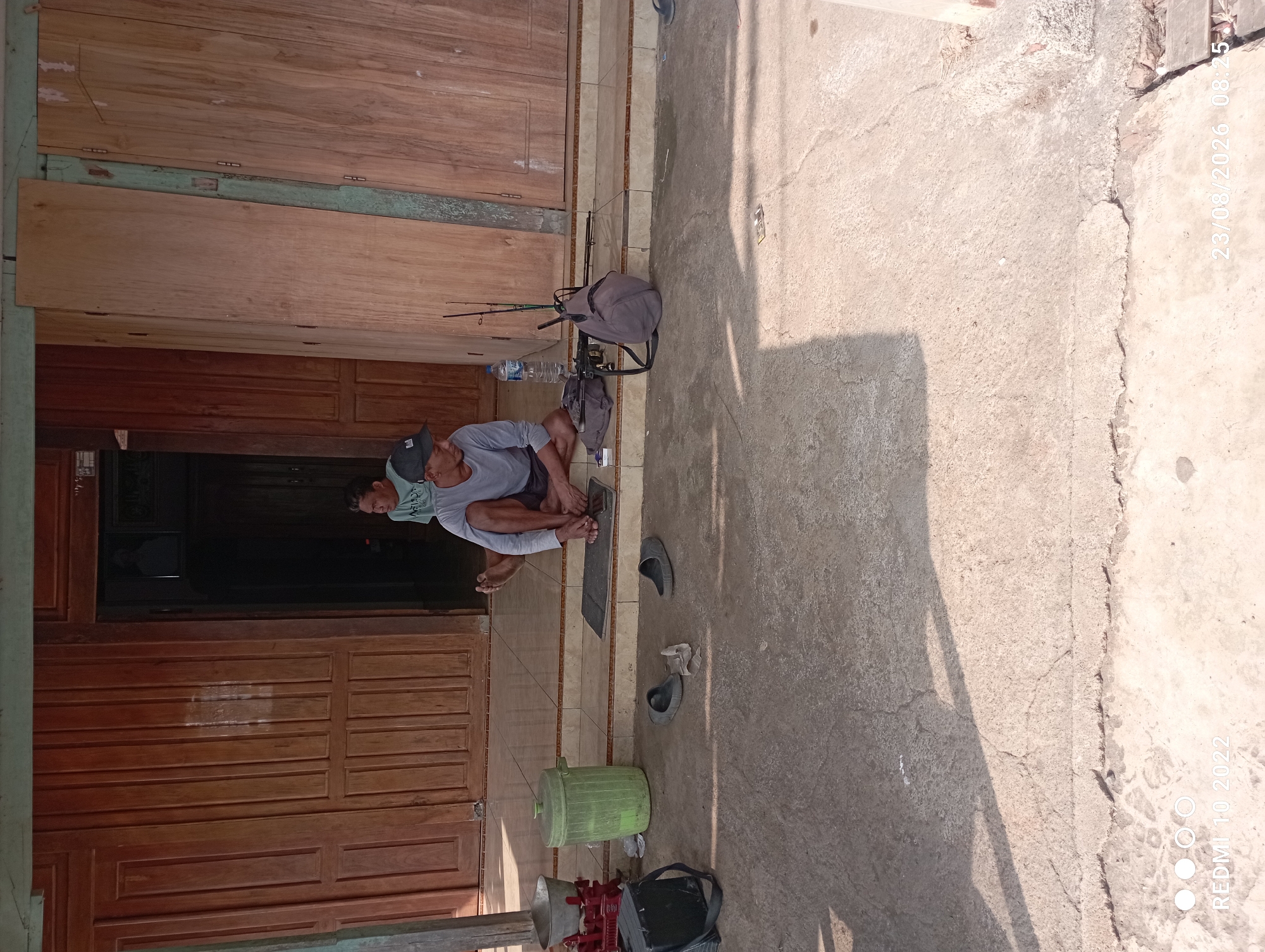 In the city of Dukuhseti, Indonesia, two men are sitting on the steps of a wooden building. The man in the foreground is wearing a gray long-sleeved shirt and dark shorts, with a black baseball cap on his head. He is seated cross-legged, sharpening a stick with a knife. Next to him are two fishing rods, a gray backpack, a blue water bottle, and a bag. Two pairs of black flip-flops are on the ground in front of him, along with some crumpled paper. To his left, a green trash can and a red-and-silver scale are visible. A second man, wearing a light blue t-shirt, sits behind and to the left of the first man, appearing to observe him. The building has large double wooden doors. The ground is paved with concrete. The timestamp on the image indicates it was taken on August 23, 2026, at 08:25.