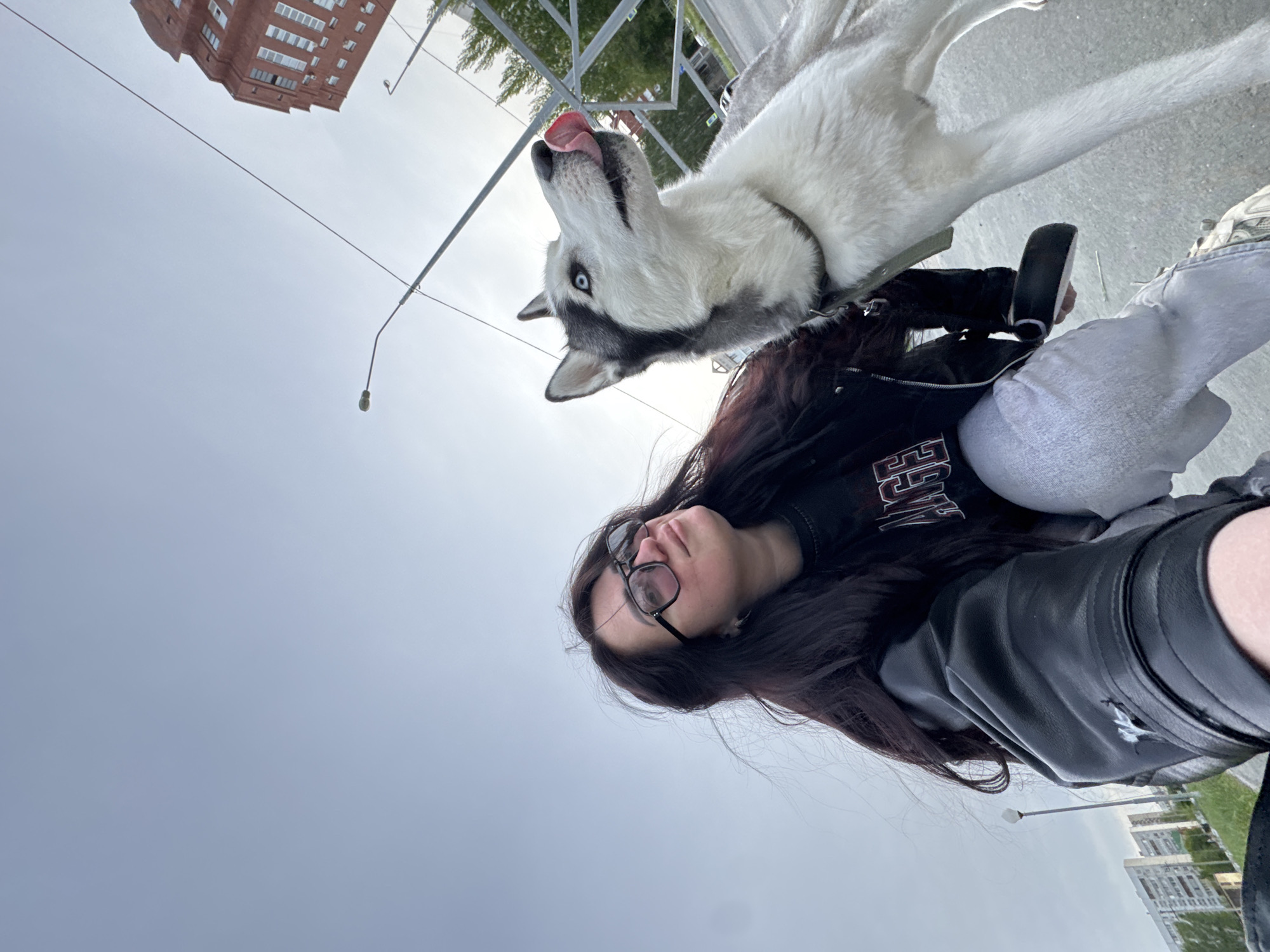 A woman stands in a city environment with a dog. She has dark hair and is wearing glasses, a black jacket, and light-colored jeans. The dog, a husky with black and white fur, is on a leash and is licking its nose. In the background, there is a building and street lights under a cloudy sky. The specific location within the city is unclear.