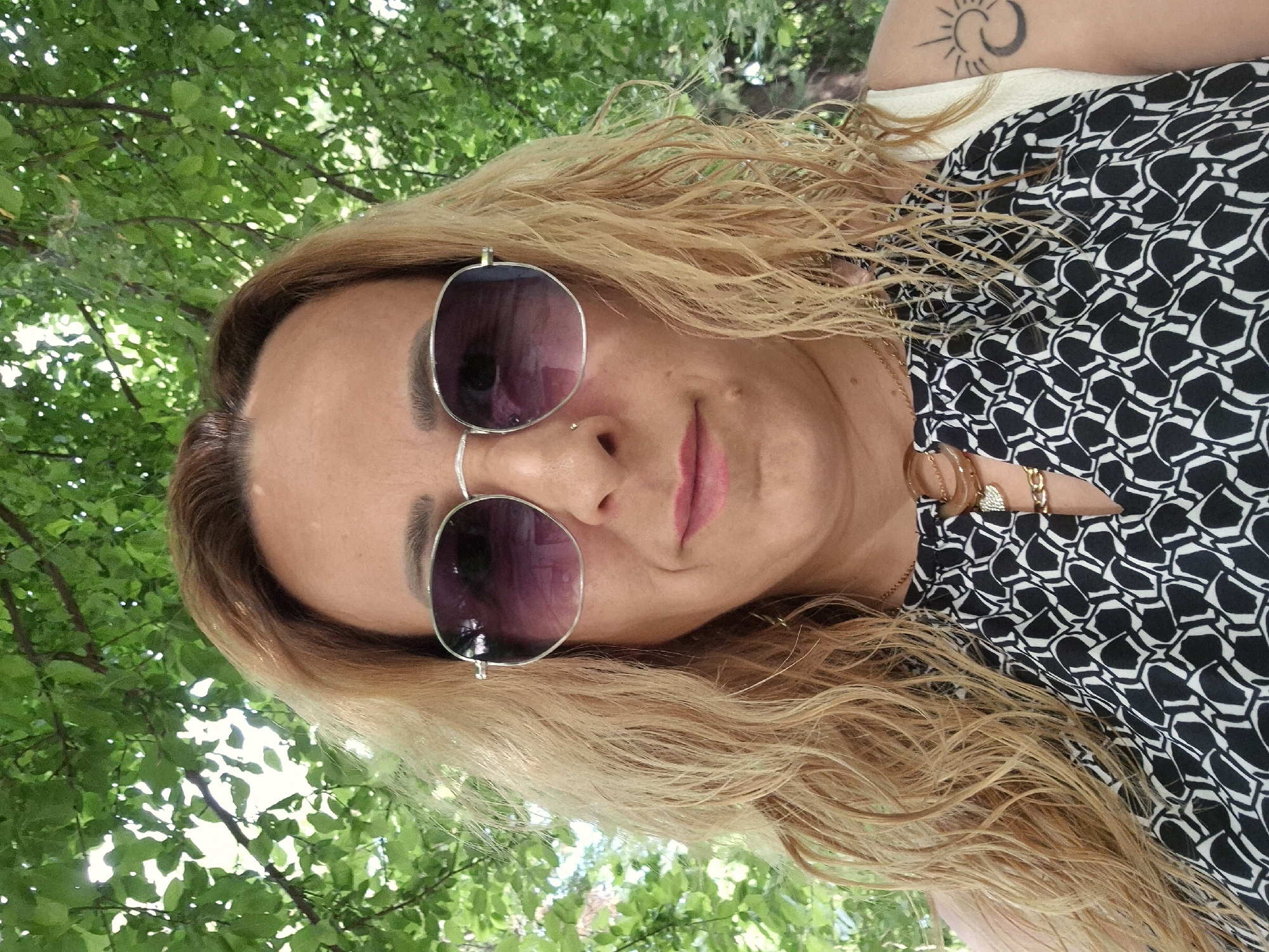 A woman poses for a selfie outdoors. She wears sunglasses, a black and white patterned top, and several gold necklaces. Her hair is blonde and wavy. On her left upper arm, a tattoo is visible. The background consists of lush green foliage, possibly from trees.