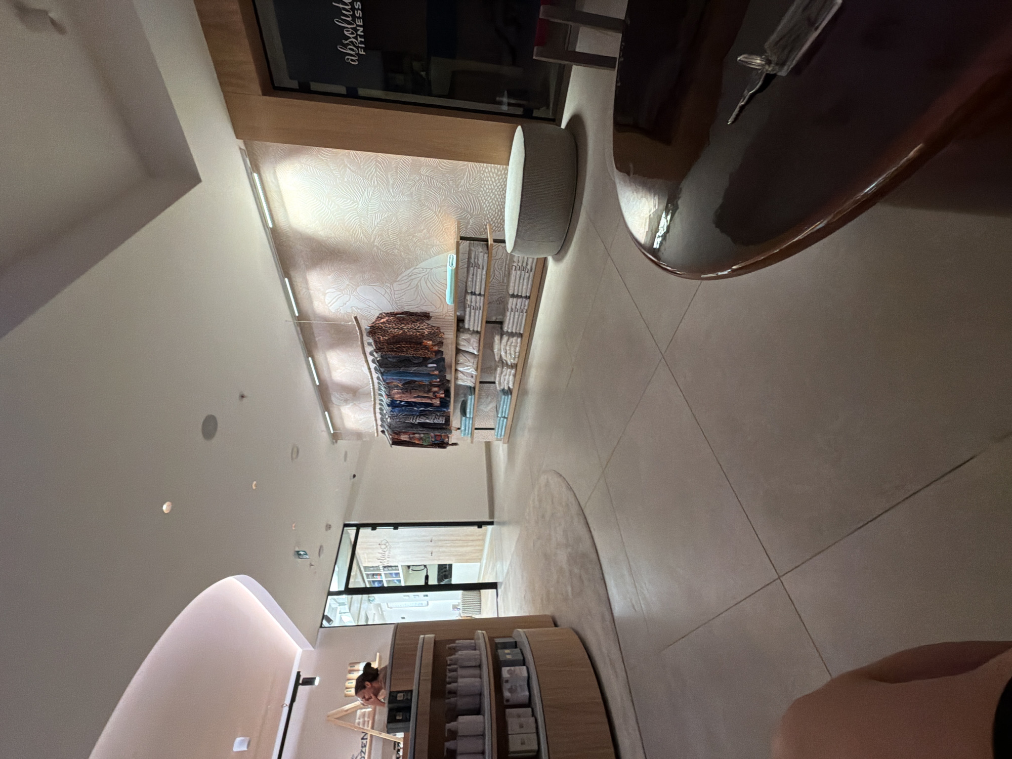 The image shows the interior of a store in Boreti, Montenegro. The store appears to be a retail establishment selling clothing and possibly other lifestyle products. In the foreground, a round, dark wooden table with a glossy finish is visible, with a set of keys resting on its surface. The floor is tiled in a neutral, stone-like pattern.
Further into the store, a curved wooden display unit is stocked with various bottles and boxes, likely toiletries or beauty products. To the left, a woman is partially visible behind the counter, appearing to be interacting with a product.
A pathway leads further into the store, where a rack of patterned clothing hangs on display. Below the clothing, shelves are neatly stacked with folded items, possibly towels or blankets. In the background, a door with glass panels provides a glimpse into another area of the store, revealing more shelving and products.
A light-colored, oval-shaped ottoman or pouf sits on the floor near the clothing display. The walls of the store feature a subtle, textured wallpaper with a leaf-like pattern, illuminated by track lighting. A sign visible on the glass door reads "absolute FITNESS". The overall aesthetic of the store is modern and clean, with warm lighting and natural materials.