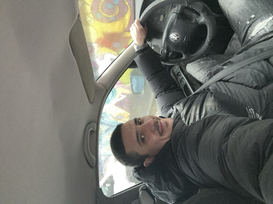 The image captures a young man, likely in his 20s or 30s, seated in the driver's seat of a car, looking directly at the camera. He has short dark hair, a mustache, and a light beard, and his expression is neutral to slightly smiling. He is dressed in a dark, puffy winter jacket and blue jeans, with a rip visible on the knee of his right pant leg. A silver-toned watch adorns his left wrist, and his hand rests on the steering wheel, which clearly displays the "Volkswagen" logo.
The setting is inside the car, with parts of the light-colored car ceiling, a-pillar, and a grab handle visible. Through the car's wet windows, a vibrant and blurred background of swirling colors, predominantly yellow, orange, blue, and green, is visible. This wet and colorful exterior strongly suggests the car is currently inside an automatic car wash, with water being sprayed on the exterior. The activity appears to be the man posing for a photo or selfie while his car is being washed. The specific location context is Bischheim, France. The time of day is not discernible, but the scene is brightly lit, consistent with the artificial lighting found in a car wash.