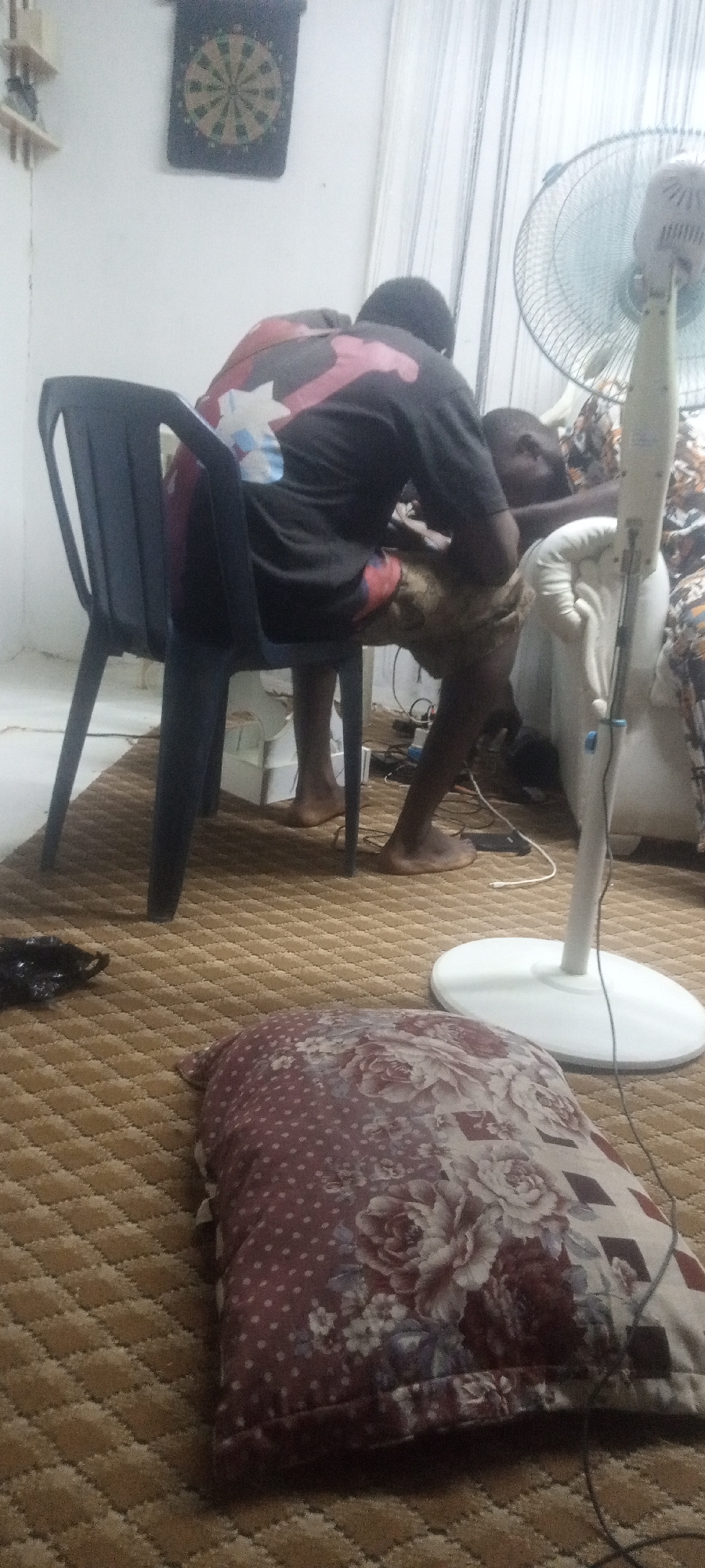 A low-angle, medium shot depicts a scene within a room. A person wearing a black t-shirt and patterned shorts is seated on a black plastic chair. They appear to be leaning over another individual who is partially visible. Beside them is a white standing fan. The foreground features a pillow with a floral and polka dot pattern resting on a brown patterned carpet. On the wall, a dartboard is visible. The location appears to be a domestic interior in the city of Unknown, Unknown.