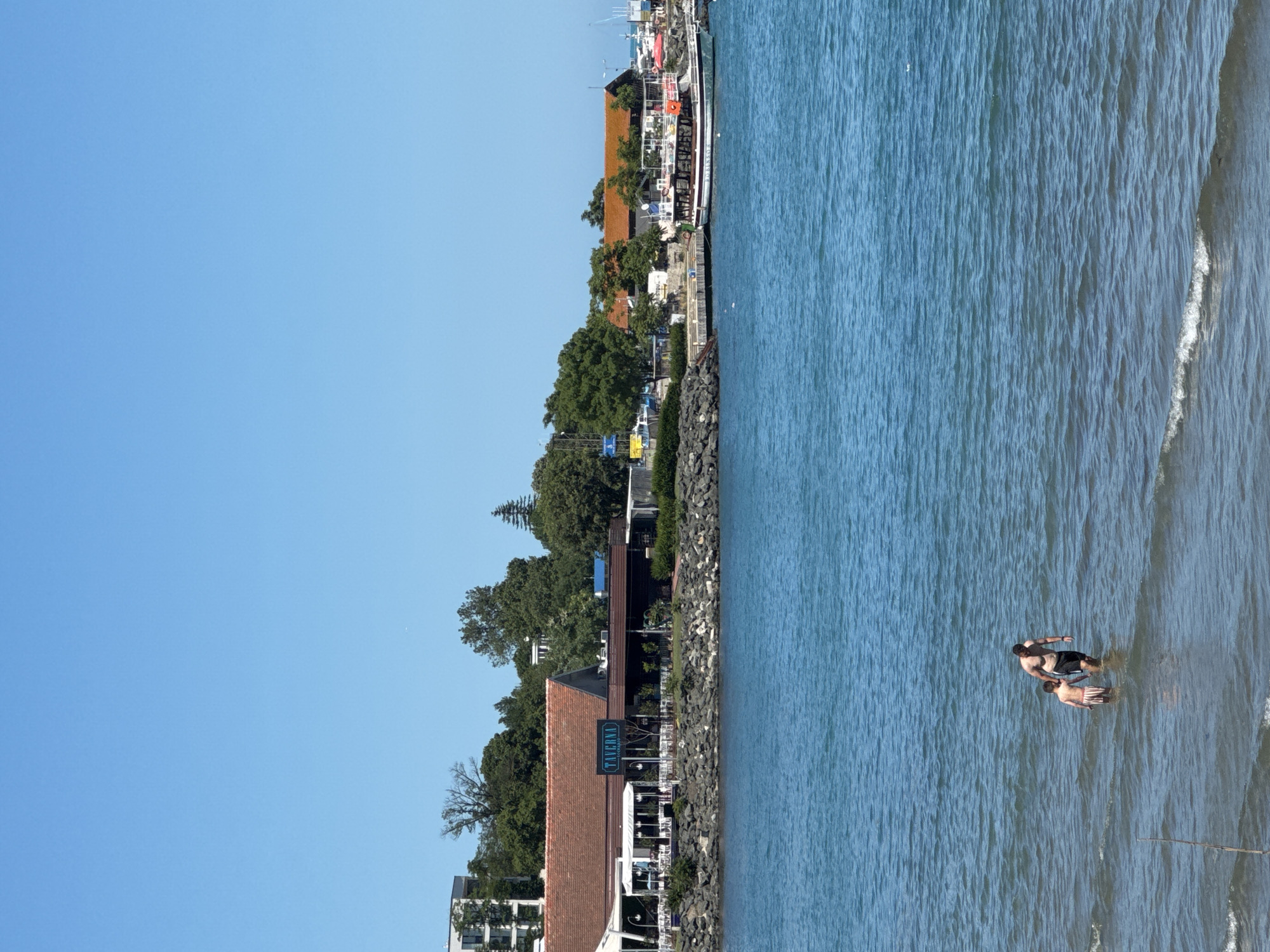 In Tsarevo, Bulgaria, two individuals, a man and a child, are wading in the calm blue sea under a clear sky. The man, wearing dark swim shorts, walks beside the child, who is wearing striped swim trunks. They are in shallow water, with small waves lapping at their legs.
In the background, a picturesque waterfront scene unfolds. A long, low-slung building with a reddish-brown tiled roof runs along the shore, identified by a neon sign reading "TAVERNA". This establishment appears to be a restaurant or cafe, with outdoor seating visible under awnings. Beyond the taverna, more buildings and lush green trees are visible, suggesting a developed coastal area. Further out in the water, several boats are moored, including a white and green passenger boat with dark trim, and other smaller vessels. The sea is a vibrant blue, reflecting the bright, sunny weather. The shore is lined with a rugged rock wall.
