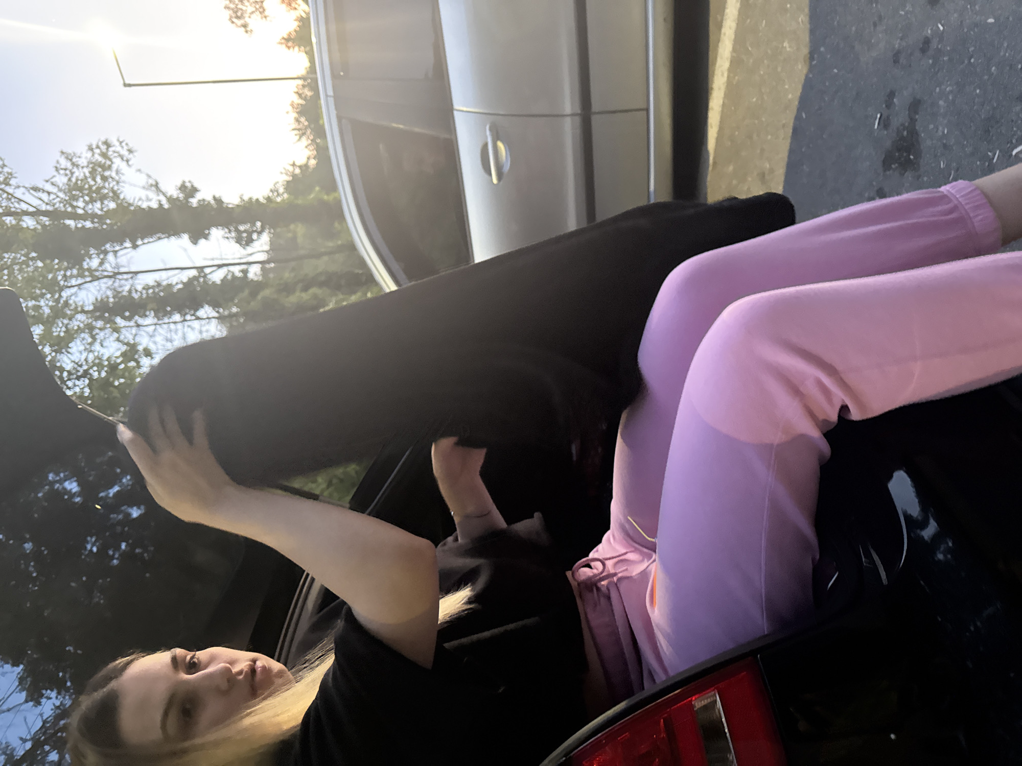 A woman with blond hair is positioned inside the trunk of a black car, wearing pink sweatpants and a black shirt. She appears to be either entering or exiting the trunk. Another car is parked nearby. Trees are visible in the background. The location is not discernible as Pleven, Bulgaria based on the image.