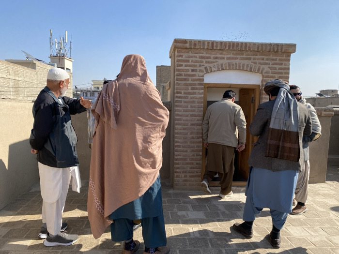 The scene depicts a rooftop in Kandahar, Afghanistan, under a clear blue sky. Five individuals are present on a tiled surface. On the far left, an older male with a white beard and skullcap, wearing a dark jacket and white traditional clothing, gestures with his right hand towards the center of the frame. Adjacent to him, a person fully covered in a light brown chador or large shawl, over dark blue clothing, stands with their back to the viewer. In the central right, a single-story brick structure with a light-colored arched doorway and an open wooden door is visible. A male in a light brown jacket and brown traditional clothing is entering this structure, seen from behind. To his right, another male in a grey patterned jacket and a grey and brown striped shawl draped over his shoulders stands, observing the entrance. A fifth male, partially obscured, stands behind the shawl-wearing individual, wearing sunglasses. In the background, distant buildings of light-colored brick are visible, and a communication tower with multiple antennas is present on a rooftop to the far left. A flock of small, dark birds is in flight above the brick structure. Strong sunlight casts clear shadows on the tiled rooftop.