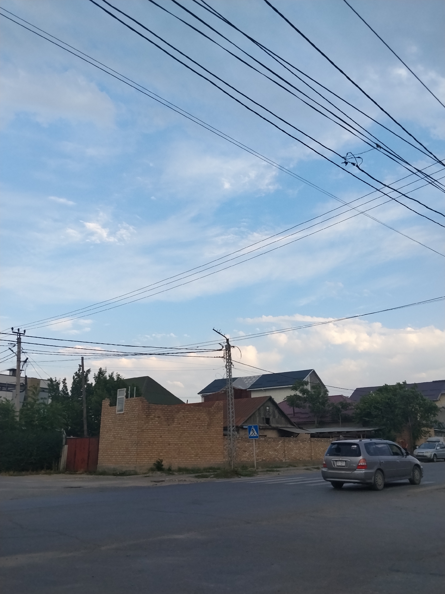 A street scene with cars and buildings under a cloudy sky. A gray minivan is parked on the right side of the road, facing away from the camera. Behind it, a silver sedan is visible. Buildings with various rooflines and brick walls line the background. Power lines crisscross the sky, with some poles visible on the left. A pedestrian crossing sign is posted on the left side of the road. The sky is a mix of blue and white clouds, suggesting it is daytime, likely late afternoon or early evening due to the lighting. The overall scene appears to be a residential or mixed-use urban area. No people are clearly visible. There is some text on the license plate of the minivan, but it is not legible.