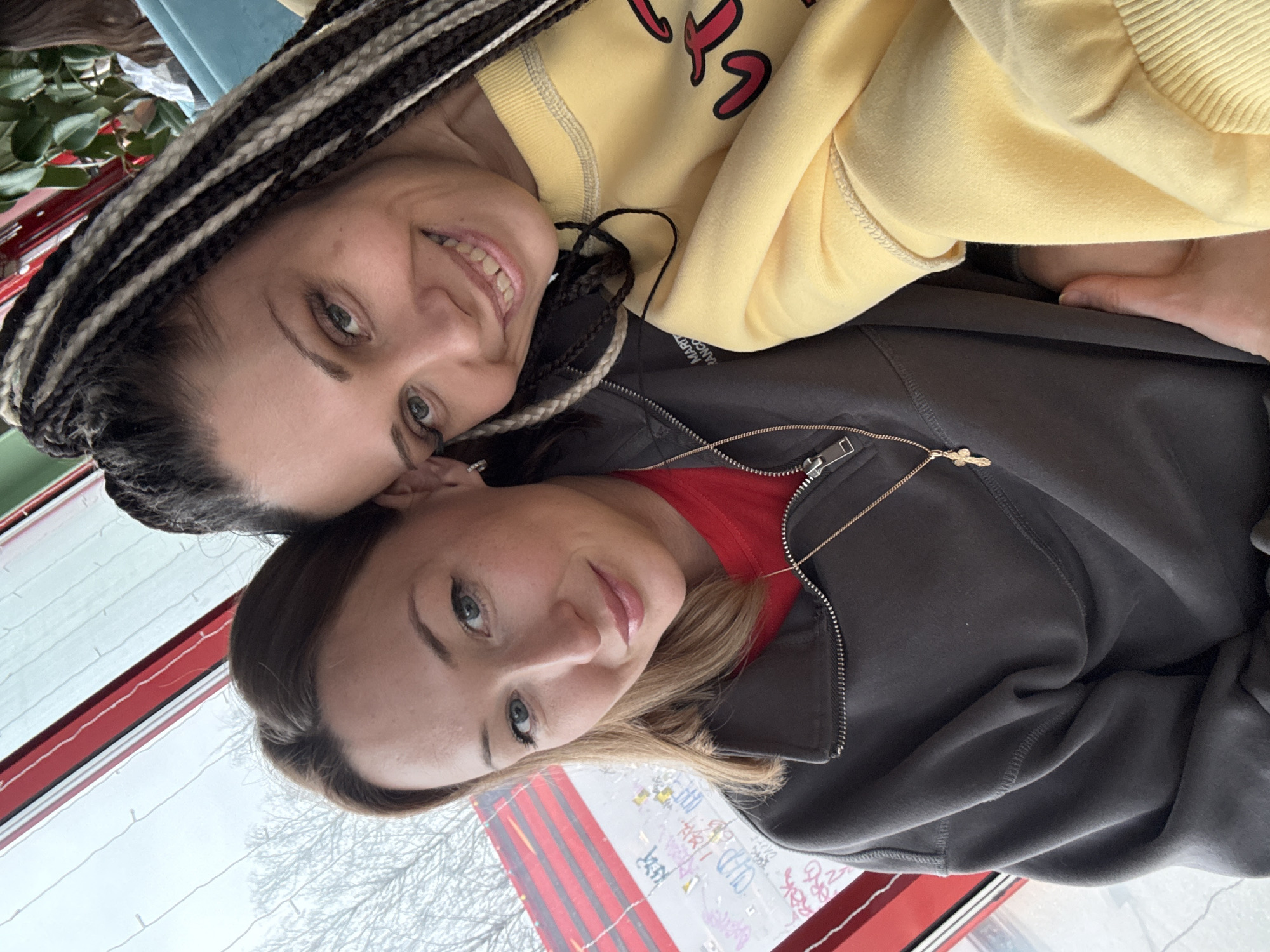 Two women are posing closely together for a selfie. The woman on the left has dark hair braided with lighter strands and is wearing a yellow sweatshirt. The woman on the right has shoulder-length blonde hair and is wearing a gray jacket over a red top. A small gold cross hangs from a necklace around her neck. They are indoors, with a window behind them. The window has a red frame, and outside, a blurred view of trees and possibly holiday decorations is visible. The location is in the city of Unknown, Unknown.
